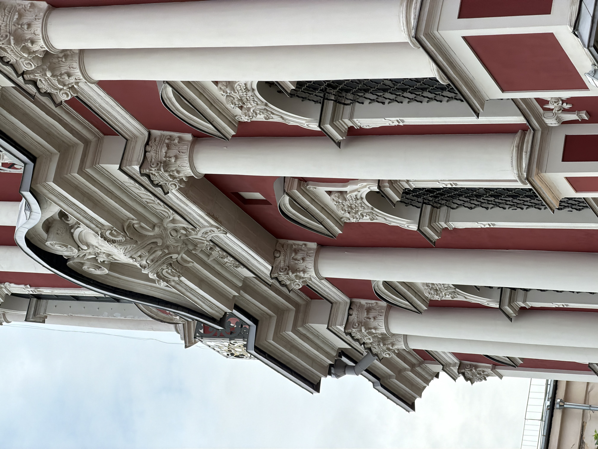 The image shows an architectural detail, most likely from a building in Moscow, Russia. The view is an upward angle, focusing on a series of columns and the ornate design of the building's facade. The columns are white, and the wall behind them is painted a deep red with white architectural trim. The craftsmanship appears to be detailed with elaborate ornamentation at the top of each column. The sky is visible in the lower portion of the image, suggesting an exterior shot. A gutter system is visible in the foreground.