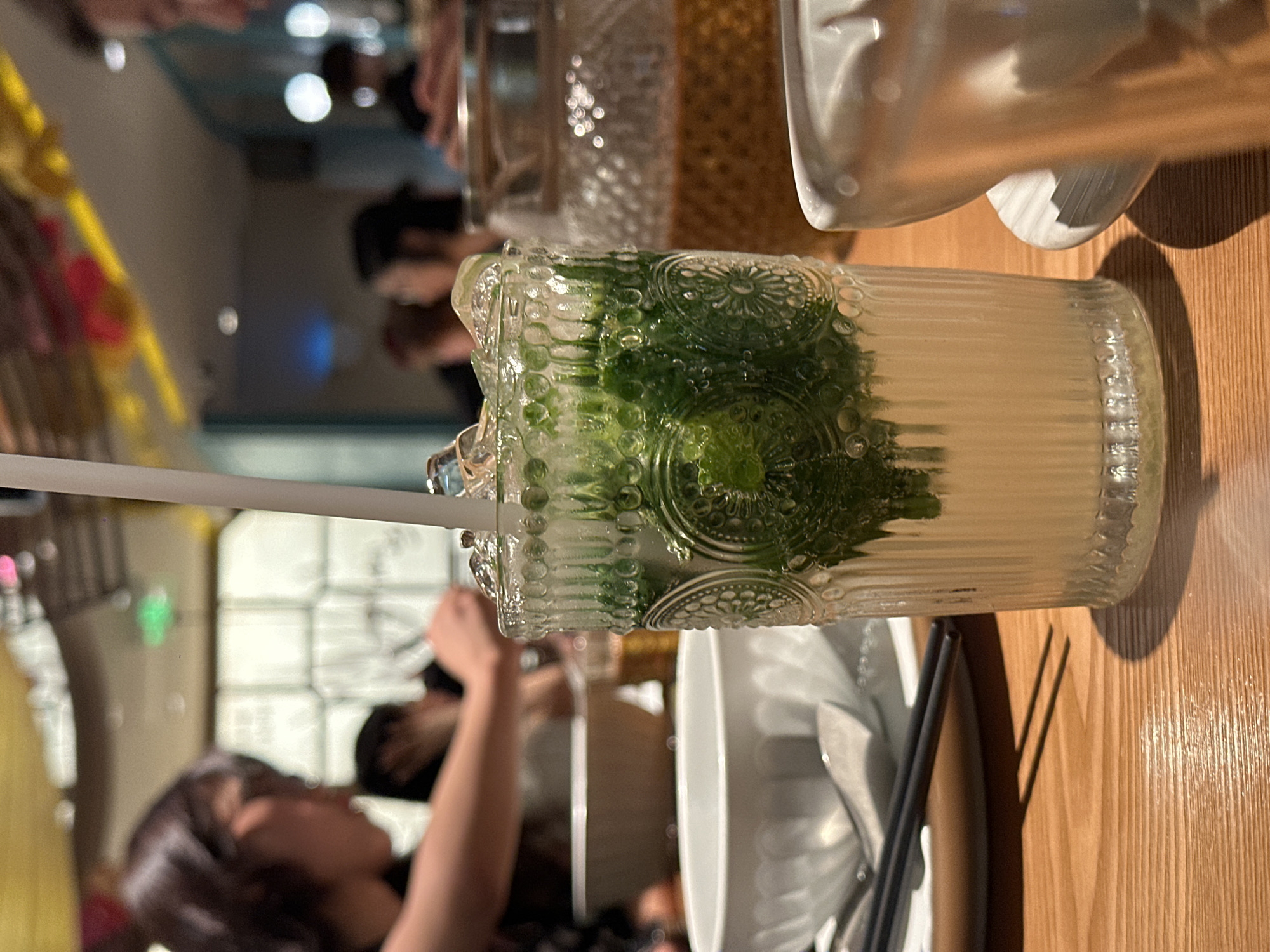 The image depicts a close-up of a refreshing drink in a decorative glass, placed on a wooden table. The drink appears to be a mojito or a similar cocktail, with crushed ice, mint leaves, and a pale yellow liquid, topped with a white straw. The glass has an embossed, ornate pattern. In the blurred background, several people are visible, some engaged in dining activities. There's a large glass pitcher behind the drink, filled with a light brown liquid. The overall setting suggests a restaurant or bar in Futian District, China, characterized by warm ambient lighting and what appears to be decorative elements on the ceiling. The focus is primarily on the drink, with the background elements providing context of a social gathering.