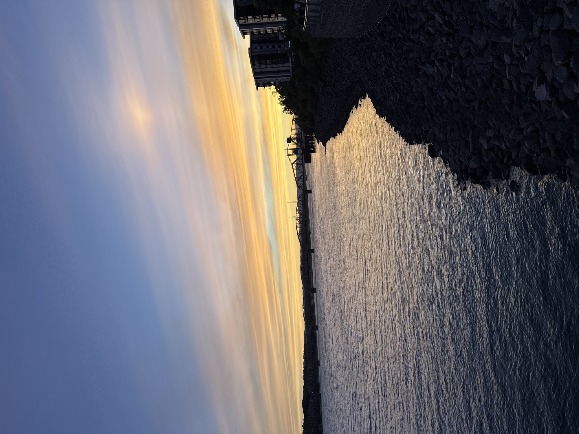 This is a photograph of a waterfront scene at dusk in Vancouver, Washington, USA. The sky is filled with vibrant streaks of orange and yellow clouds, transitioning into a soft blue overhead. A large body of water, likely the Columbia River, dominates the left side of the frame, with gentle ripples reflecting the sunset colors. The right bank of the river is lined with dark, irregular rocks. Beyond the rocky embankment, there are modern, multi-story buildings with dark facades and visible windows. A pedestrian walkway with a railing is visible along the top of the embankment, with a few silhouetted figures walking along it. In the distance, across the water, a long, multi-span bridge with a dark metal structure is visible, with a smaller, older bridge structure closer to the viewer's side. The overall scene captures a peaceful evening ambiance with urban elements.
