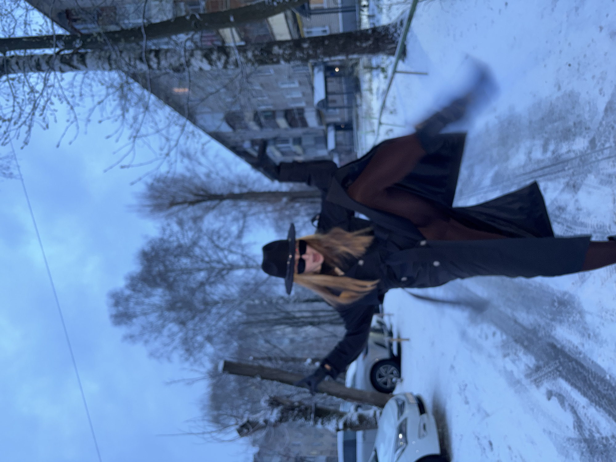 A long-haired person is captured mid-action, striking a dynamic pose on a snow-covered street in Perm, Russia. The individual, likely female, wears a black wide-brimmed hat, dark sunglasses, a long black coat, dark leggings or tights, and boots. One leg is lifted high, and the person's arms are slightly spread, with one gloved hand seemingly gripping a wooden post or railing.
The setting is an urban or suburban street during winter, characterized by significant snow cover on the ground, revealing tire tracks. In the background, multiple multi-story residential buildings are visible, typical of an urban environment. Bare trees, some with frost or snow on their branches, line the street. Two white cars are partially visible, parked on the snowy roadside. The sky is overcast and a pale blue-grey, indicating a cold, winter day with diffused light. No visible text is present in the image.