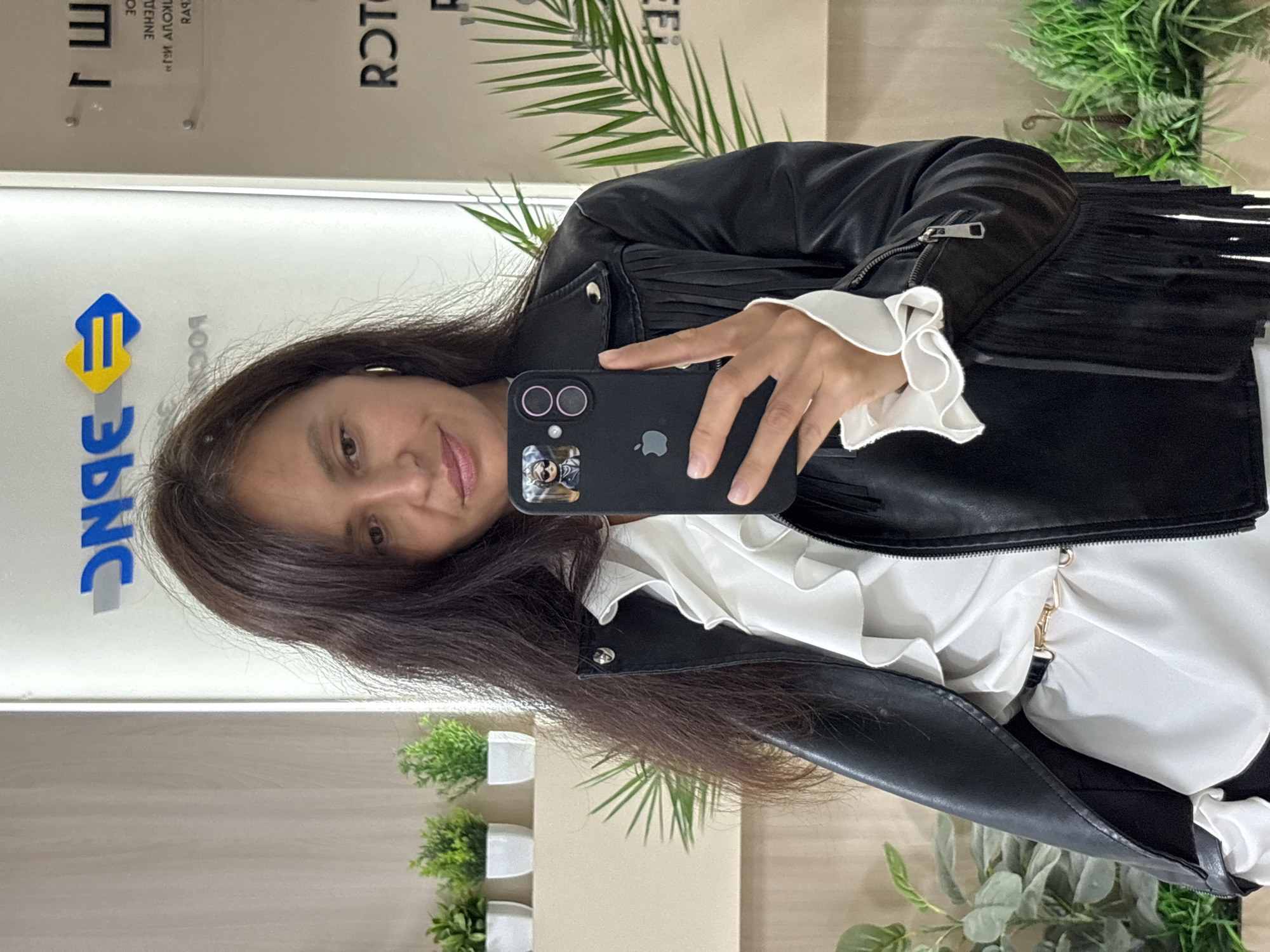 A woman is taking a selfie with a black smartphone. She is wearing a black leather jacket with fringe and a white ruffled blouse. She has long brown hair. The background appears to be an office space with plants and a sign with Russian text. The sign includes the logo of "ЭБНС." Given the presence of the logo and the city in the prompt, the location may be the ЭБНС office in Chaikovsky, Russia.