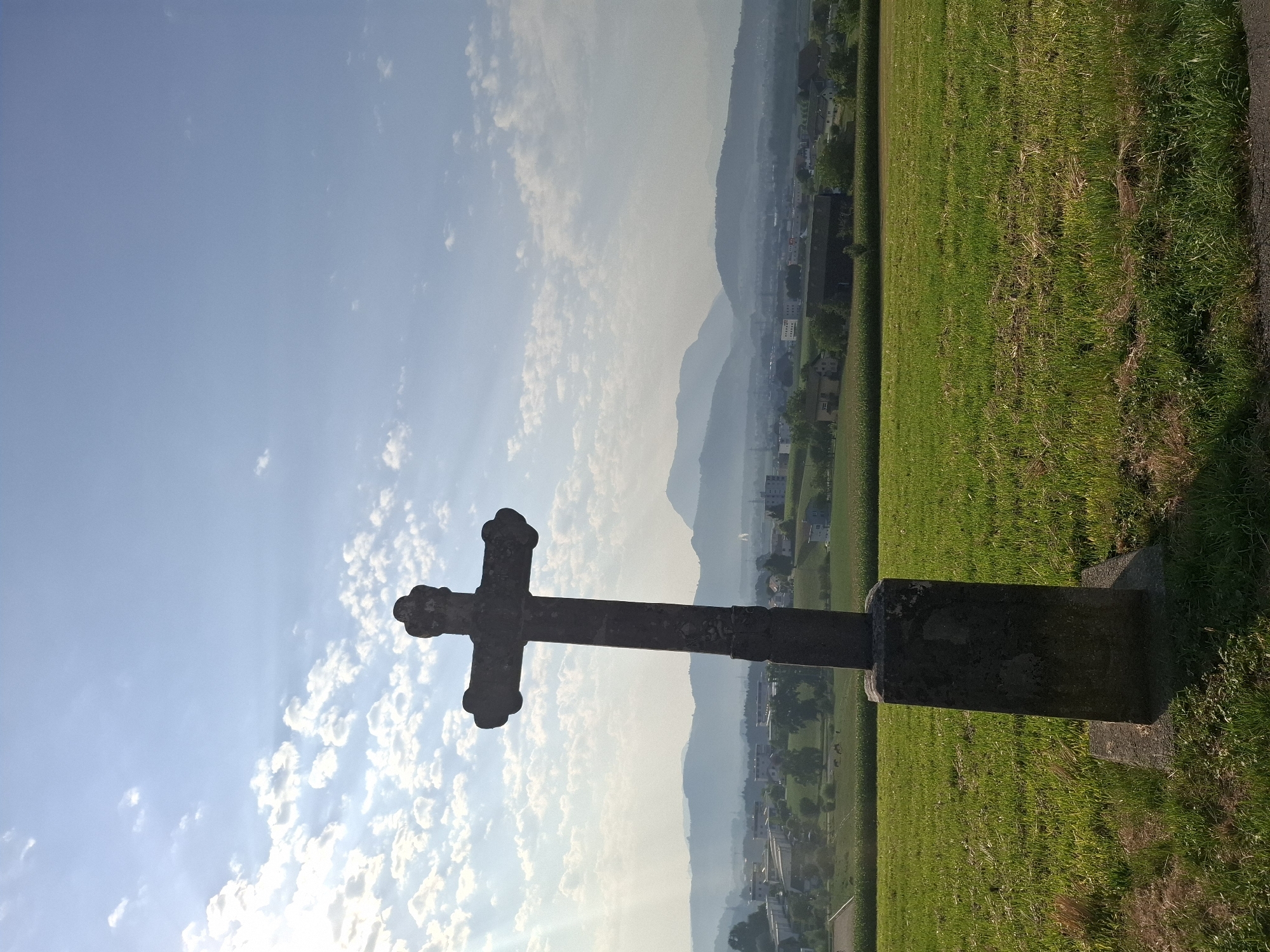 A stone cross stands in the foreground on a grassy hill, silhouetted against a bright sky. The cross is dark and weathered, with a detailed, ornate top. The base of the cross is a rough, rectangular stone structure. The hill slopes down towards a valley where a town is visible, with buildings and houses scattered amongst trees. Beyond the town, rolling hills and mountains recede into the hazy distance. The sky is blue with scattered white clouds, and sunlight streams from the upper left, casting a shadow from the cross onto the grass. The environment is rural and peaceful, with a sense of elevation and a panoramic view. There are no people visible in the image. The specific location is on a hill overlooking the municipality of Eschenbach (LU) in Switzerland.