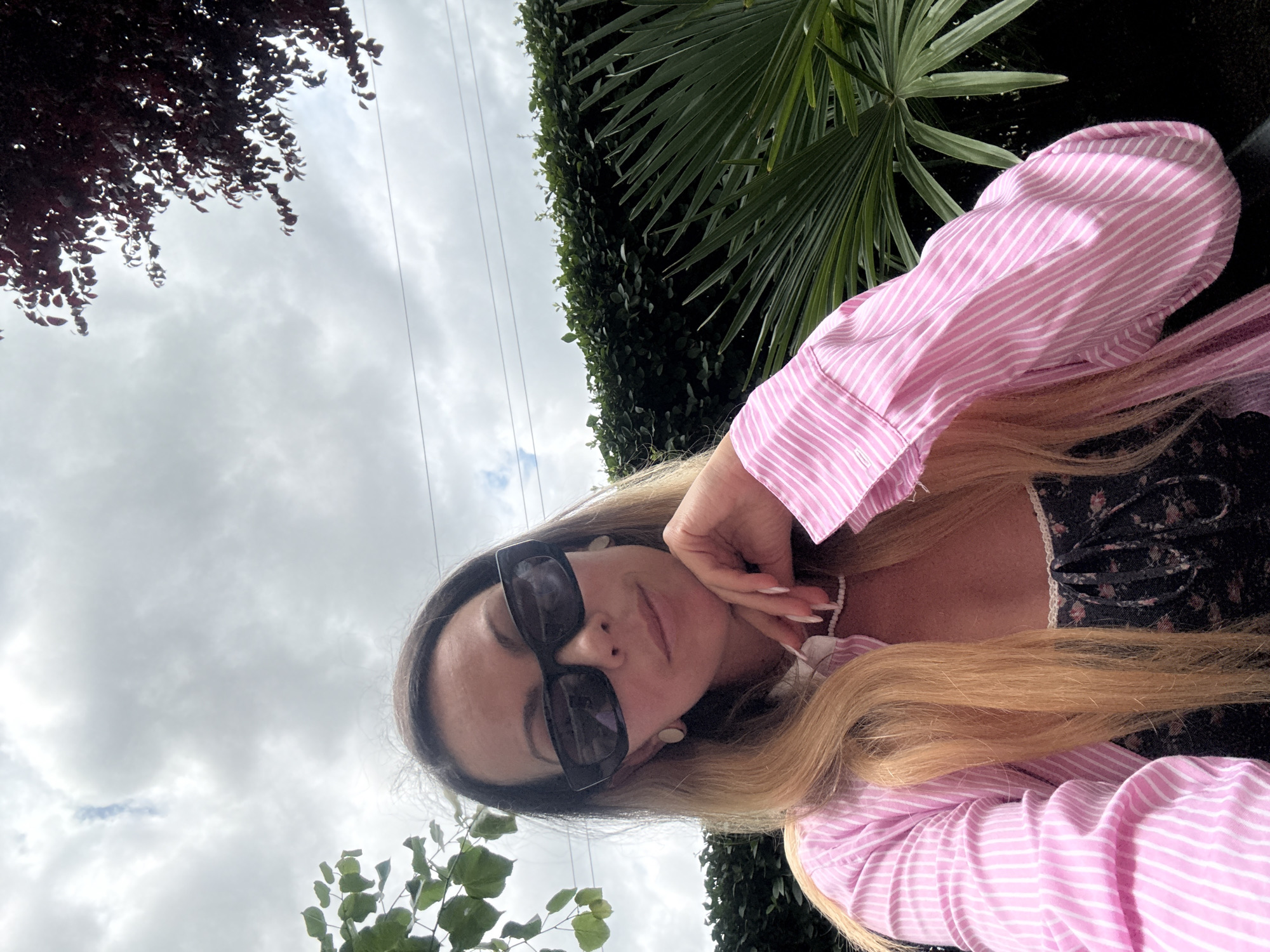 A woman with blonde hair and sunglasses is posing for a selfie outdoors. She is wearing a pink and white striped button-down shirt over a dark floral top. Her left hand is resting on her chin, and she is looking at the camera. In the background, there is a hedge, a palm frond, and a cloudy sky. The image appears to have been taken in Kosharitsa, Bulgaria.