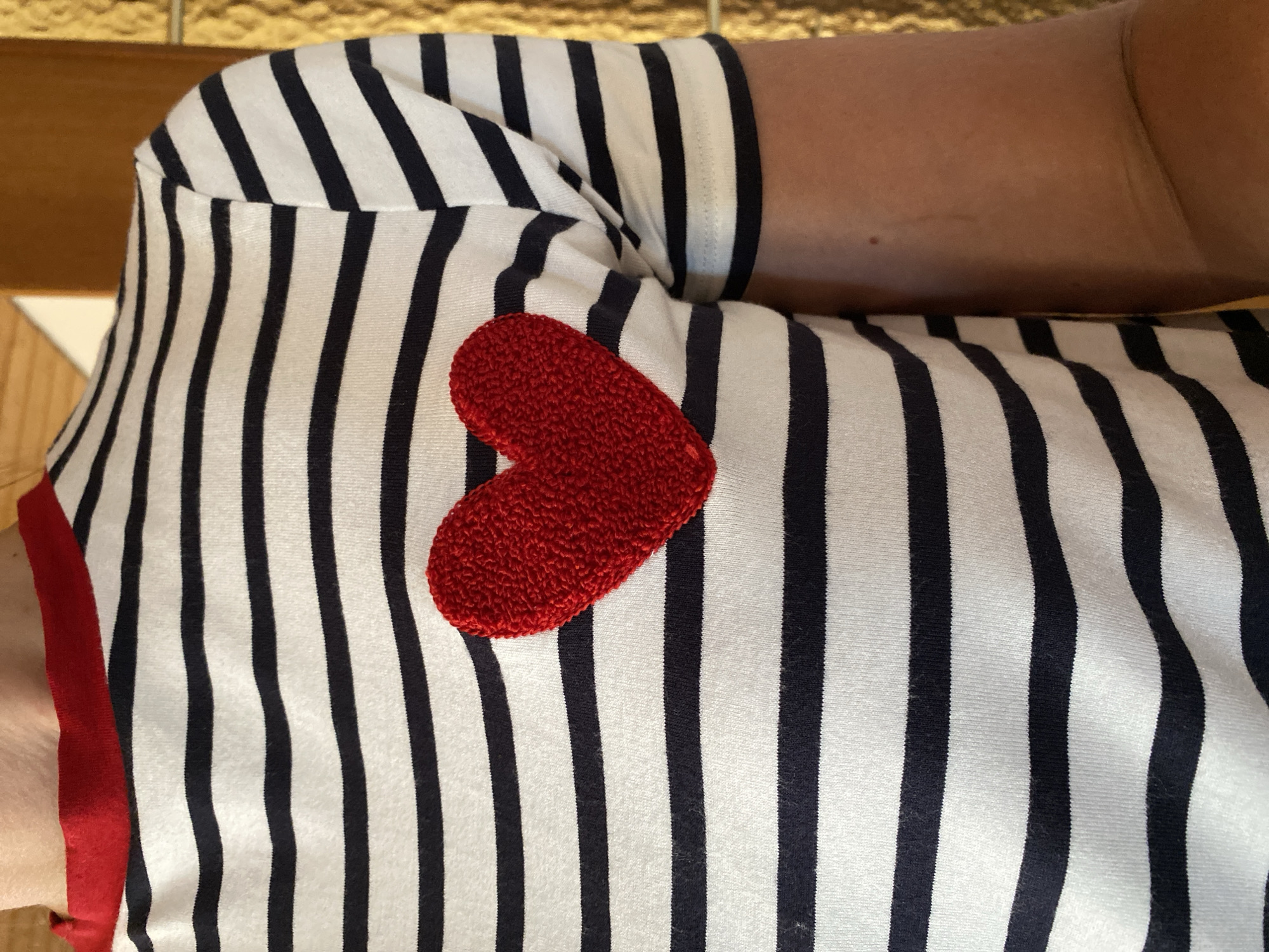 A close-up shot shows a person wearing a black and white striped t-shirt with a red collar and a red embroidered heart on the left breast. The background is a blurry golden-yellow texture, possibly a wall or fabric. The image is taken from the chest down to the arm, focusing on the t-shirt design. There is no visible connection to Flammersfeld, Germany, or any specific urban environment.