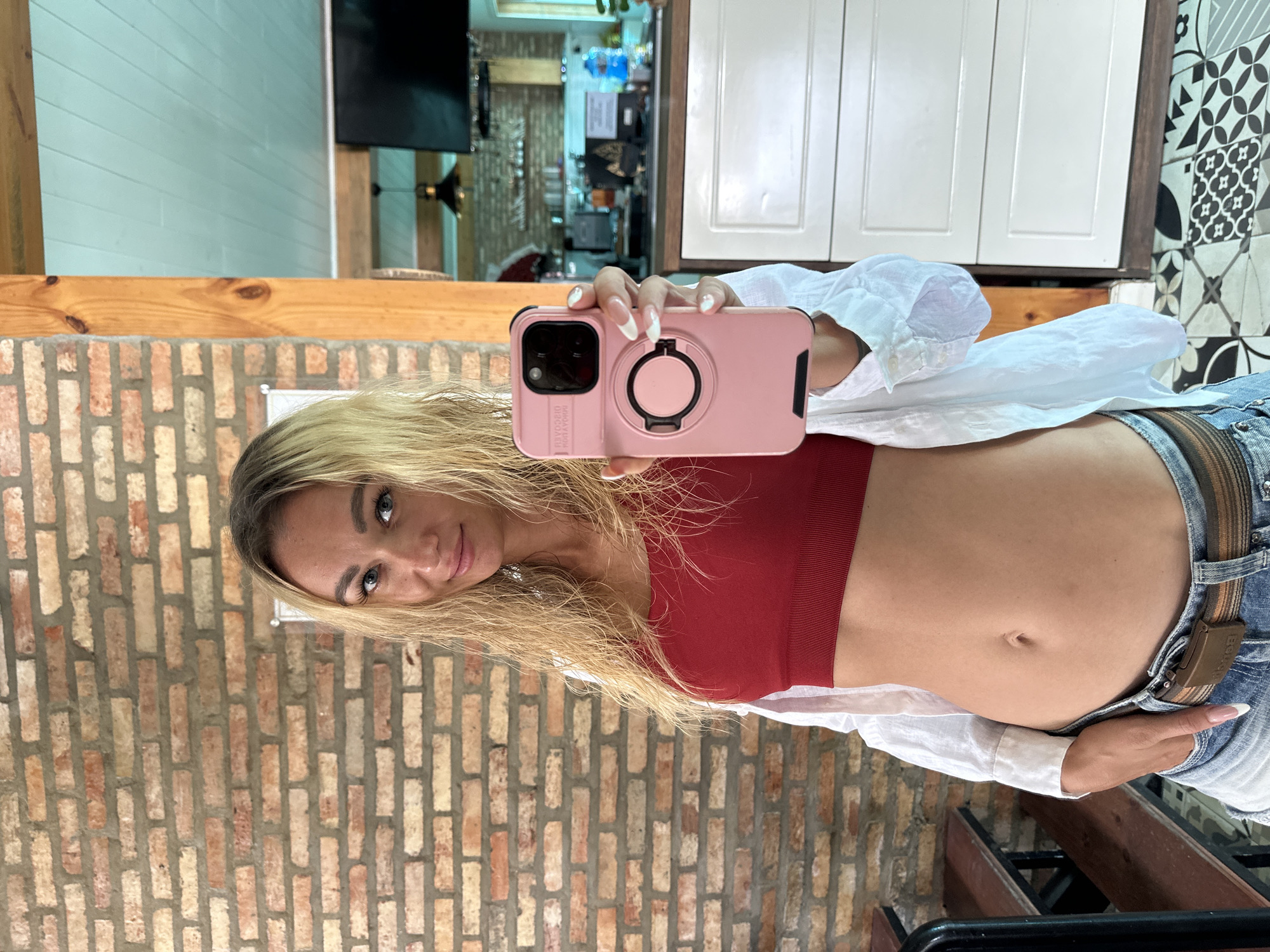 A woman with blonde hair takes a mirror selfie in a room with exposed brick walls. She is wearing a red crop top, a white button-down shirt, and denim shorts with a brown belt. Her phone, in a pink case with a ring holder, is held up to take the photo. The background shows a wall of white cabinets, a patterned tile wall, and what appears to be a wooden counter. The lighting suggests it is daytime.