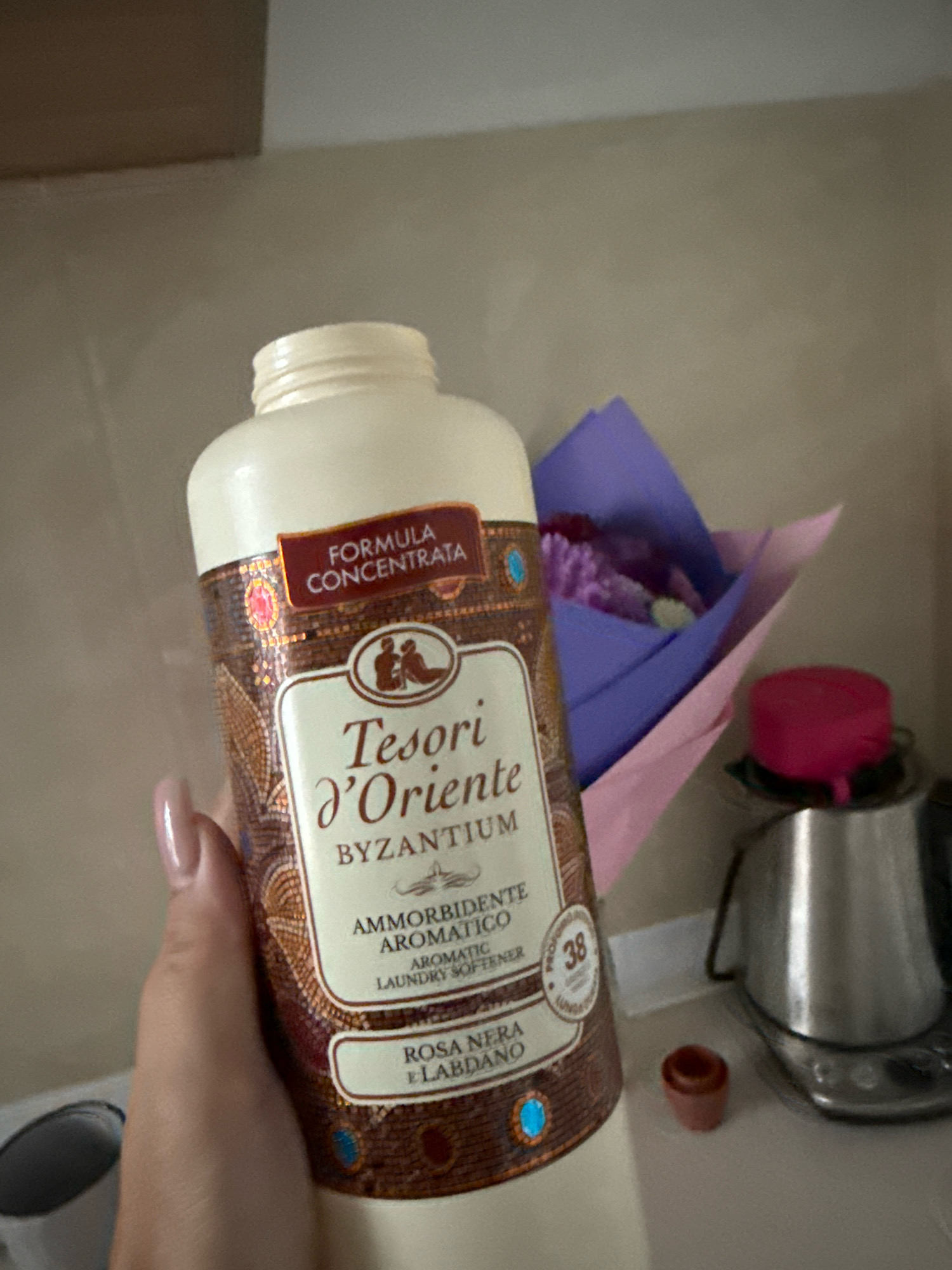 A person's hand, with manicured nails, is holding a bottle of Tesori d'Oriente Byzantium laundry softener. The bottle has a cream-colored cap and a label with intricate brown, gold, and red mosaic patterns. Text on the label indicates "FORMULA CONCENTRATA," "Tesori d'Oriente," "BYZANTIUM," "AMMORBIDENTE AROMATICO," "AROMATIC LAUNDRY SOFTENER," and "ROSA NERA E LABDANO." A small circle with the number "38" is also visible. In the background, a bouquet of flowers wrapped in purple and pink paper is placed near a silver kettle with a pink lid. The scene appears to be indoors, likely in a domestic setting, and there is no identifiable information placing it in Chișinău, Moldova.