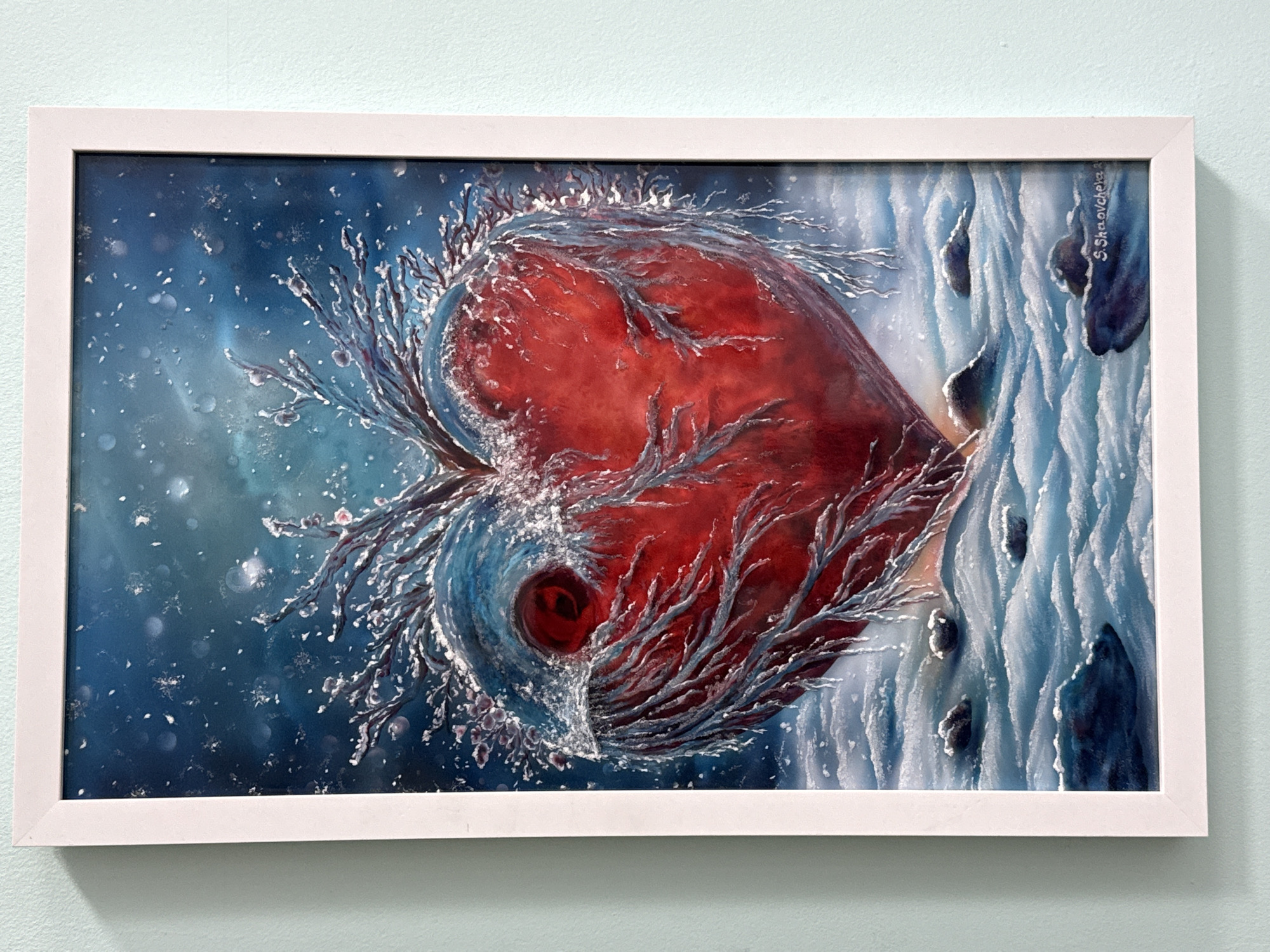 A framed painting hangs on a pale blue wall. The painting depicts a large red heart-shaped object, surrounded by icy, branch-like structures. The background is a mix of blue and white, suggesting a cold, wintry environment with falling snow. To the right of the heart, there's a section of textured white and blue that resembles a cliff or ice formation. A signature can be seen on the painting. There is no indication that this image is from the city of Burgas, Bulgaria.