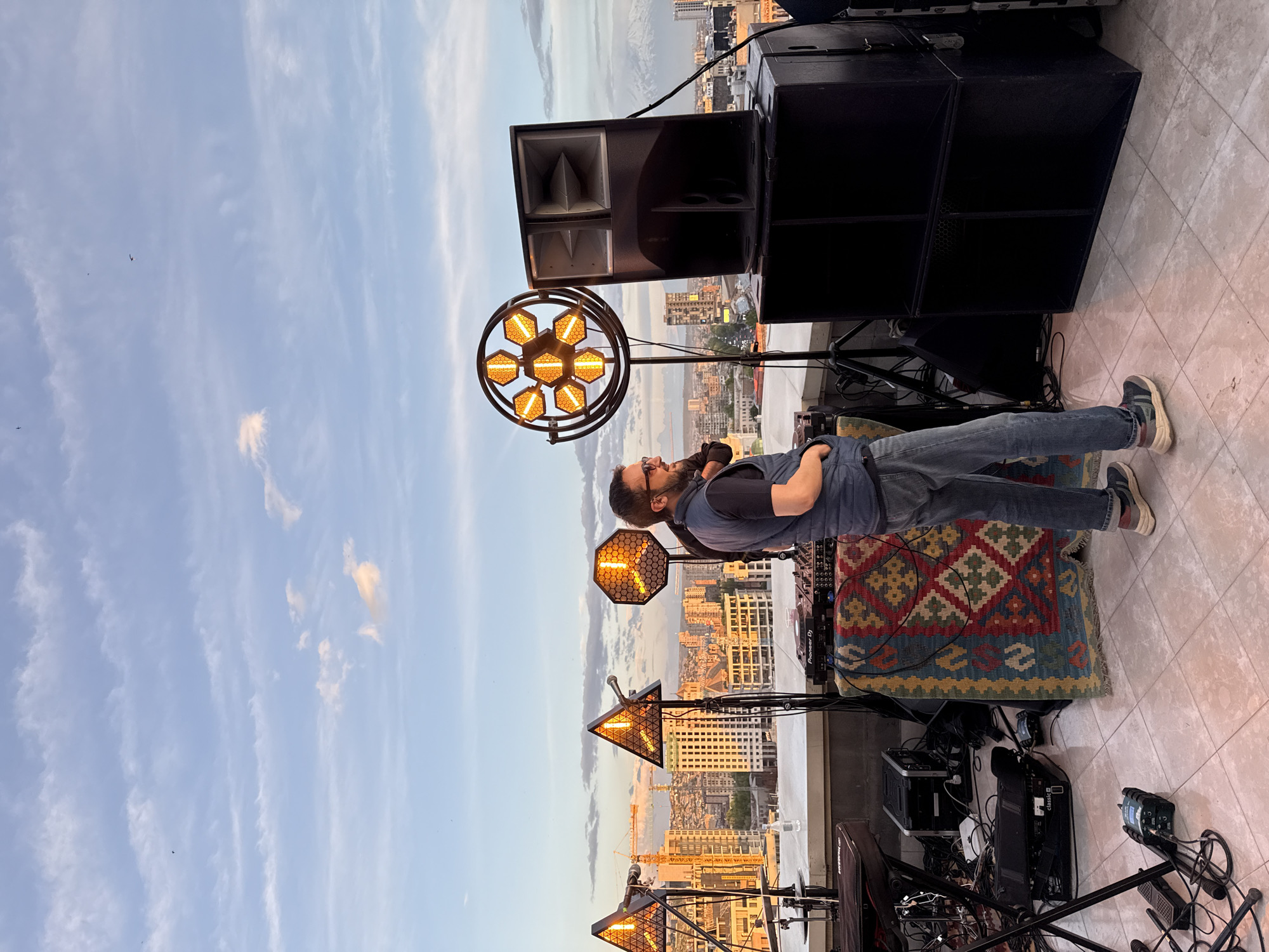 The image depicts a man standing next to DJ equipment on a rooftop in Yerevan, Armenia. The man is wearing a dark t-shirt, a denim vest, jeans, and sneakers, and he has a beard and glasses. He has his arms crossed and is looking up. The DJ setup includes speakers, a mixing console, a rug, and several stage lights. The lights are arranged in different shapes, including a circle, an octagon, and a triangle. In the background, there's a cityscape at dusk, with buildings and a crane visible. The sky is light blue with scattered clouds. The rooftop surface is tiled.