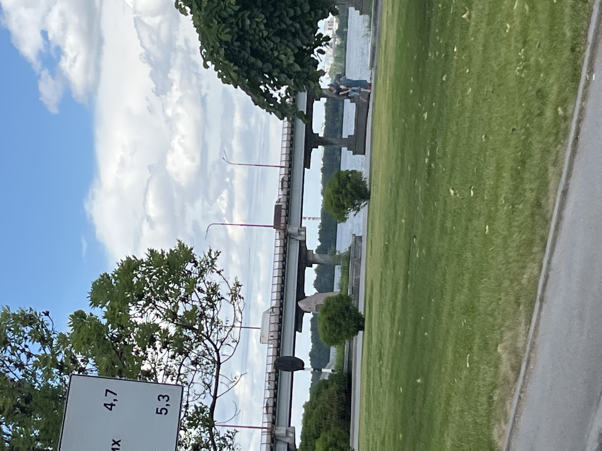 The image shows a park or waterfront area with a bridge spanning a body of water. In the foreground, there is a grassy area with some trees. A white sign with black text is partially visible, showing numbers "4,7" and "5,3", and possibly "1X" below. Further in the background, there are two people sitting on a ledge near the water, under a large tree. The bridge has multiple spans and red-colored support structures. The sky is cloudy with patches of blue. The overall scene suggests a daytime setting, possibly a mild or overcast day. The presence of the bridge and the water indicates an outdoor location in a town or city, with the provided context suggesting Dnipro, Ukraine. There are no strong emotional cues, and the activity appears to be leisure.