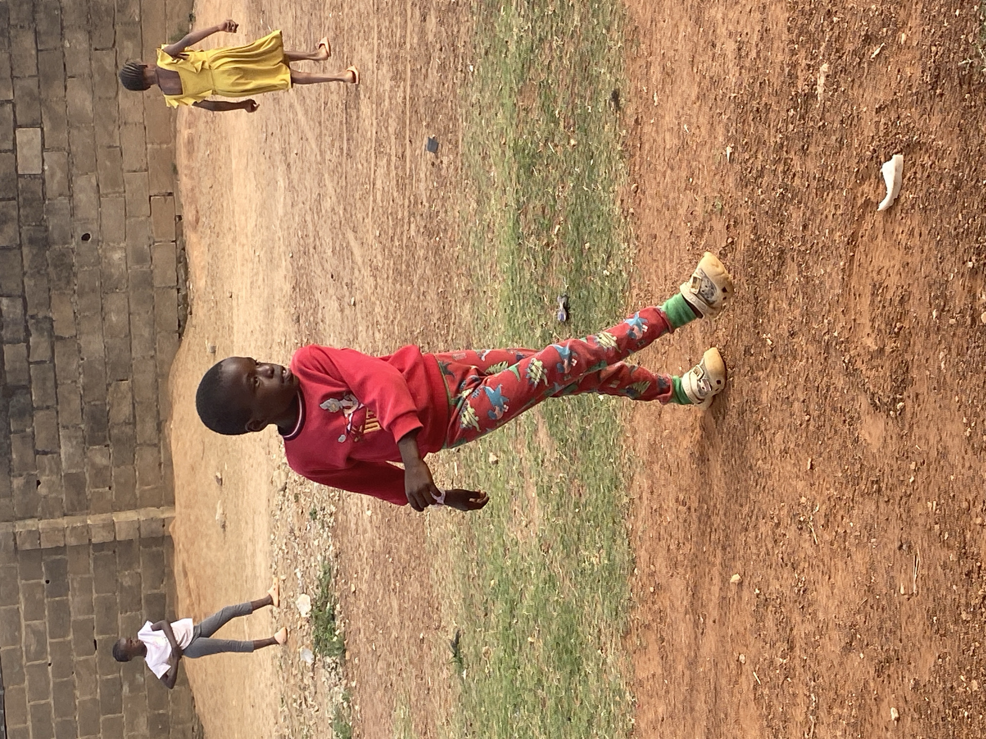 The image, taken in Bukuru, Nigeria, captures an outdoor scene featuring three children under bright daylight. The ground consists of reddish-brown earth with scattered patches of dry green grass. A wall made of concrete blocks runs along the left side of the frame.
In the foreground, a child wears a red long-sleeved shirt and colorful patterned trousers, adorned with designs in red, green, blue, and white. This child also wears light-colored shoes with green socks and is looking to their left, with an observant expression.
Further back, positioned towards the top left if the image were correctly oriented, a second child is seen wearing a bright yellow dress and light-colored sandals. This child stands facing away from the camera, looking towards the right.
In the background, towards the bottom left, a third child stands, dressed in a white t-shirt, grey trousers, and light-colored sandals. This child also appears to be facing away or slightly to the side.
The ground is dusty and uneven. A distinctive white, curved object lies on the ground to the right of the foreground child. Another small, dark object is visible near a patch of grass in the middle ground. The clear, bright lighting suggests a sunny day, likely midday or early afternoon, with no strong shadows indicating an overcast sky. There is no visible text in the image. The overall impression is a casual, everyday scene of children in their outdoor environment.