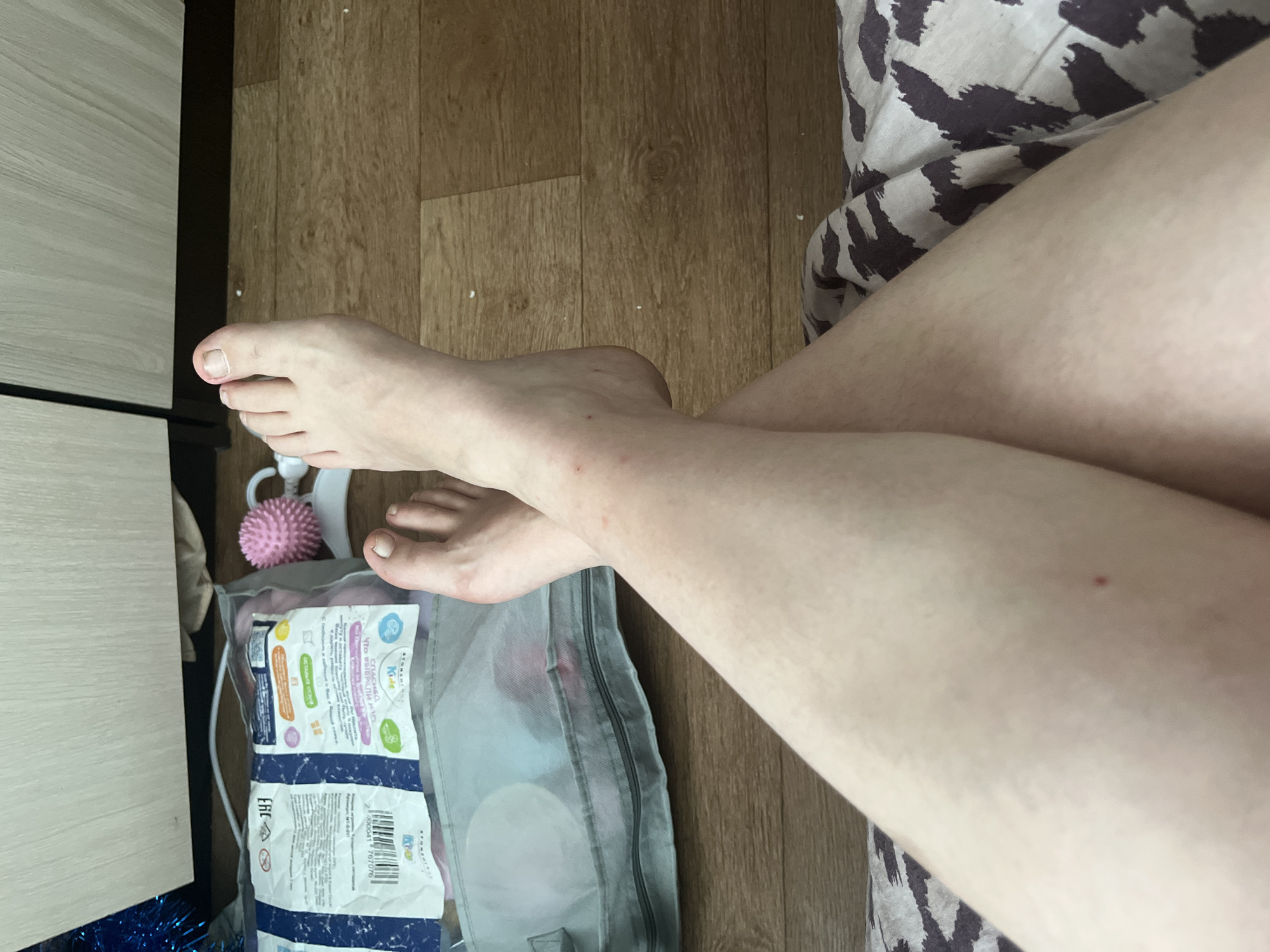 The image shows a person's legs and feet crossed, resting on a patterned fabric. In the background, there is a light-colored wooden cabinet and a floor made of wooden planks. A transparent bag containing what appear to be baby items, along with a pink spiky massage ball, is visible on the floor. The person's skin is pale, and there are a few small red marks on the legs. The lighting suggests an indoor setting, likely a domestic environment. There is no visible information in the image that would allow for the identification of a specific location within the city of Chelyabinsk, Russia.
