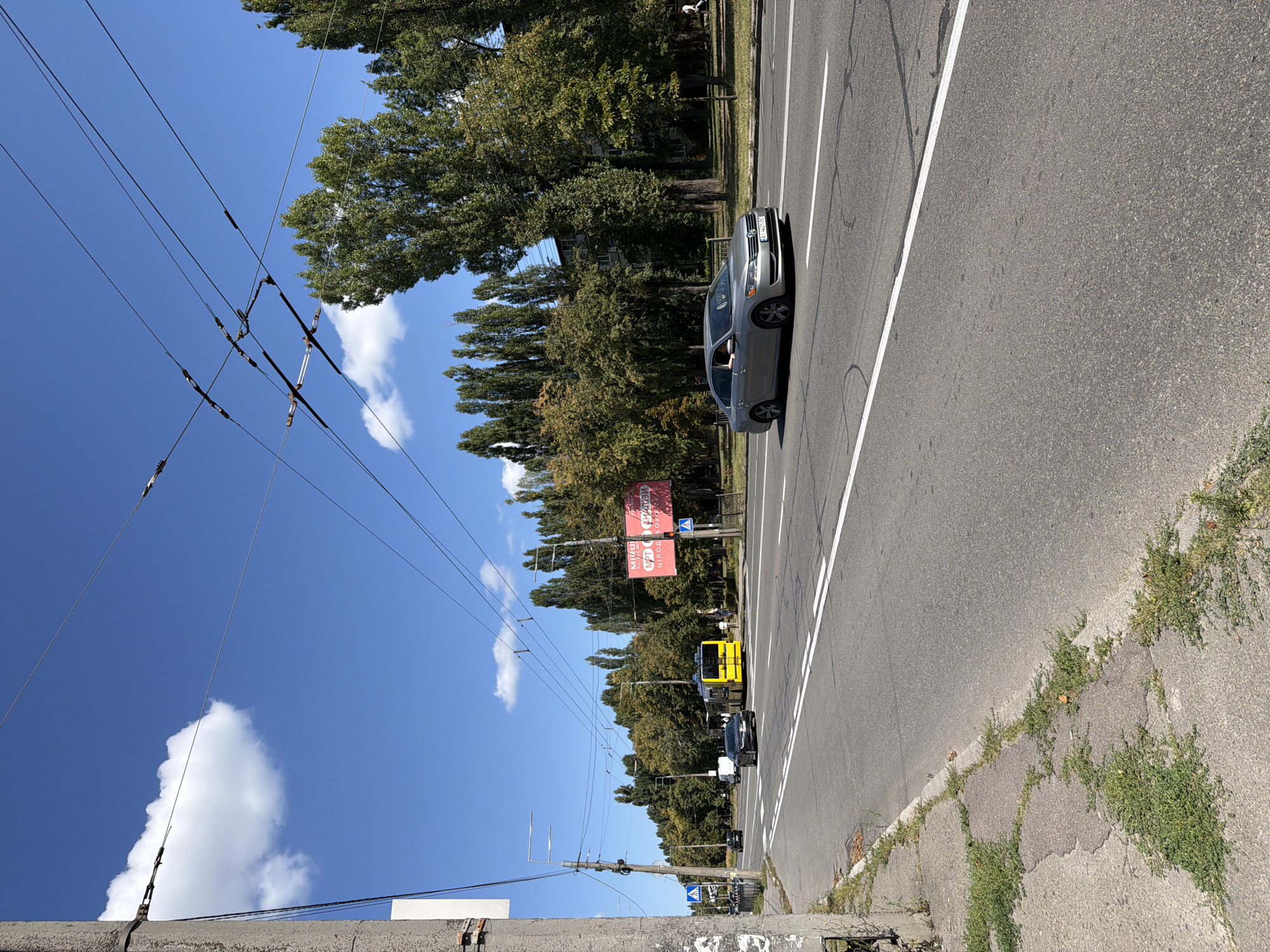 A low-angle, eye-level shot captures a sunny day in Kyiv, Ukraine, on a wide, asphalt road with visible white lane markings. Overhead, a complex network of trolleybus wires crisscrosses the bright blue sky, punctuated by a single white cloud. On the right side of the road, a line of tall, mature deciduous trees with green foliage forms a natural barrier, behind which a multi-story apartment building is partially visible. To the left of the road, a concrete sidewalk is cracked and overgrown with sparse green grass and weeds.
Several vehicles are in motion on the road. In the foreground, a silver sedan with a Ukrainian license plate (AT 4621 DC) is traveling in the right lane, with its driver visible. Further down the road, a yellow trolleybus is also moving in the right lane. A black sedan is in the left lane, and a white van is further ahead in the right lane. On the right side of the road, a large red billboard with Cyrillic text is mounted on a pole. Traffic lights are visible, with one red light illuminated. Pedestrians are sparsely scattered, with one individual walking on the sidewalk on the far right. Streetlights and utility poles are present along the side of the road. The overall scene depicts a typical urban streetscape on a clear day.