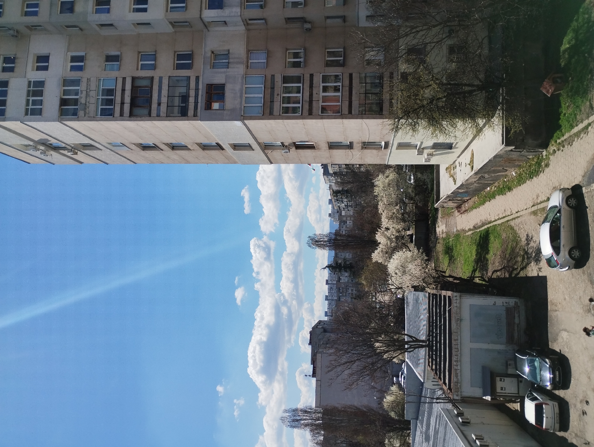 The image depicts an urban scene, likely in Sofia, Bulgaria, characterized by high-rise buildings and vehicles. The sky is blue with scattered white clouds. A tall, grey, multi-story building dominates the upper left of the frame, featuring numerous rectangular windows. Adjacent to this building is an area with trees, blossoming trees, and grassy patches. Further down, there are lower buildings, potentially garages, and parked cars. There are three visible parked cars. A pedestrian walkway bordered by greenery is visible at the edge of the frame. The overall impression is one of a densely populated urban environment with some green spaces.