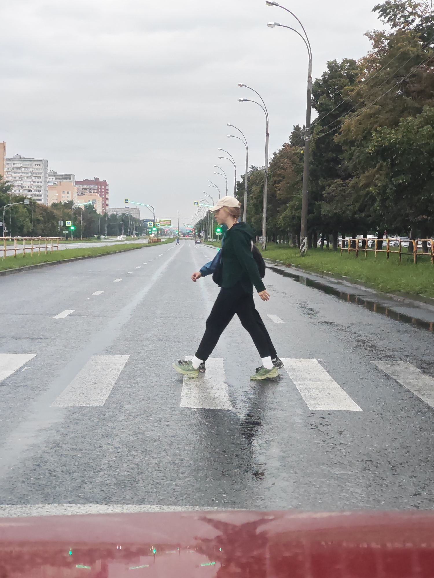 An individual wearing a beige baseball cap, a dark green hooded sweatshirt, black pants, and light green athletic shoes is crossing a wet, marked crosswalk on a city street. The street has multiple lanes and is lined with trees on the right side, with apartment buildings visible in the distance on the left. Streetlights with curved arms are positioned along the side of the road, and traffic lights are visible in the background. The ground is reflective, indicating it has recently rained or is currently raining. The lower foreground of the image shows the top of a red vehicle, possibly a car, with reflections of green lights. The sky is overcast.