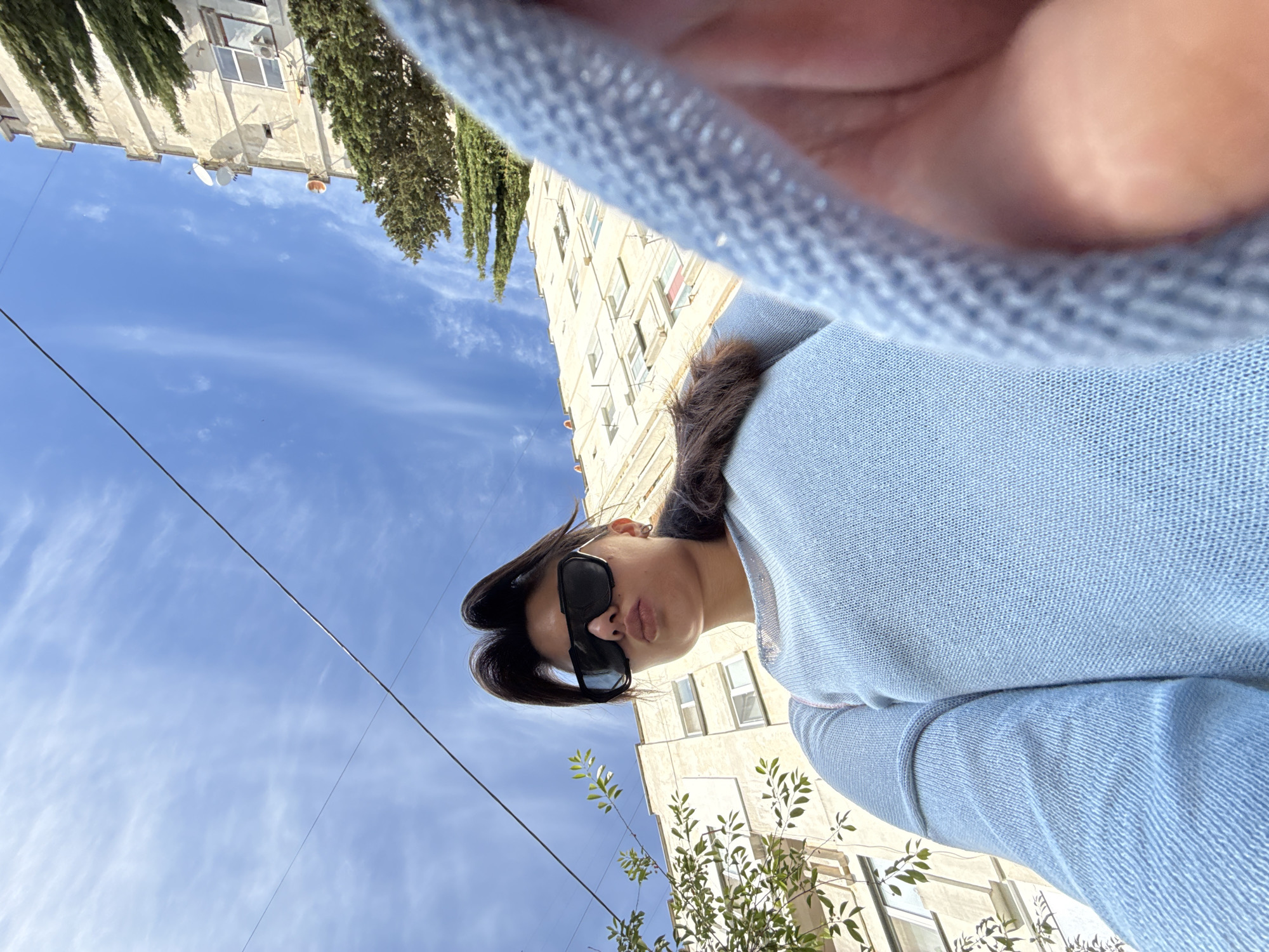 A low-angle, outdoor, waist-up selfie shows a young woman with dark hair pulled into a ponytail and styled upwards, wearing large, angular black sunglasses and a light blue knit sweater. Her lips are pursed. She is positioned in the foreground, with a pale, multi-story building and a clear blue sky with wispy white clouds filling the background. A power line runs diagonally across the upper portion of the image. Some trees with green foliage are visible to the left of the building. A blurry portion of the woman's arm, wearing a similar light blue knitted garment, is in the lower right corner, partially obscuring the scene.