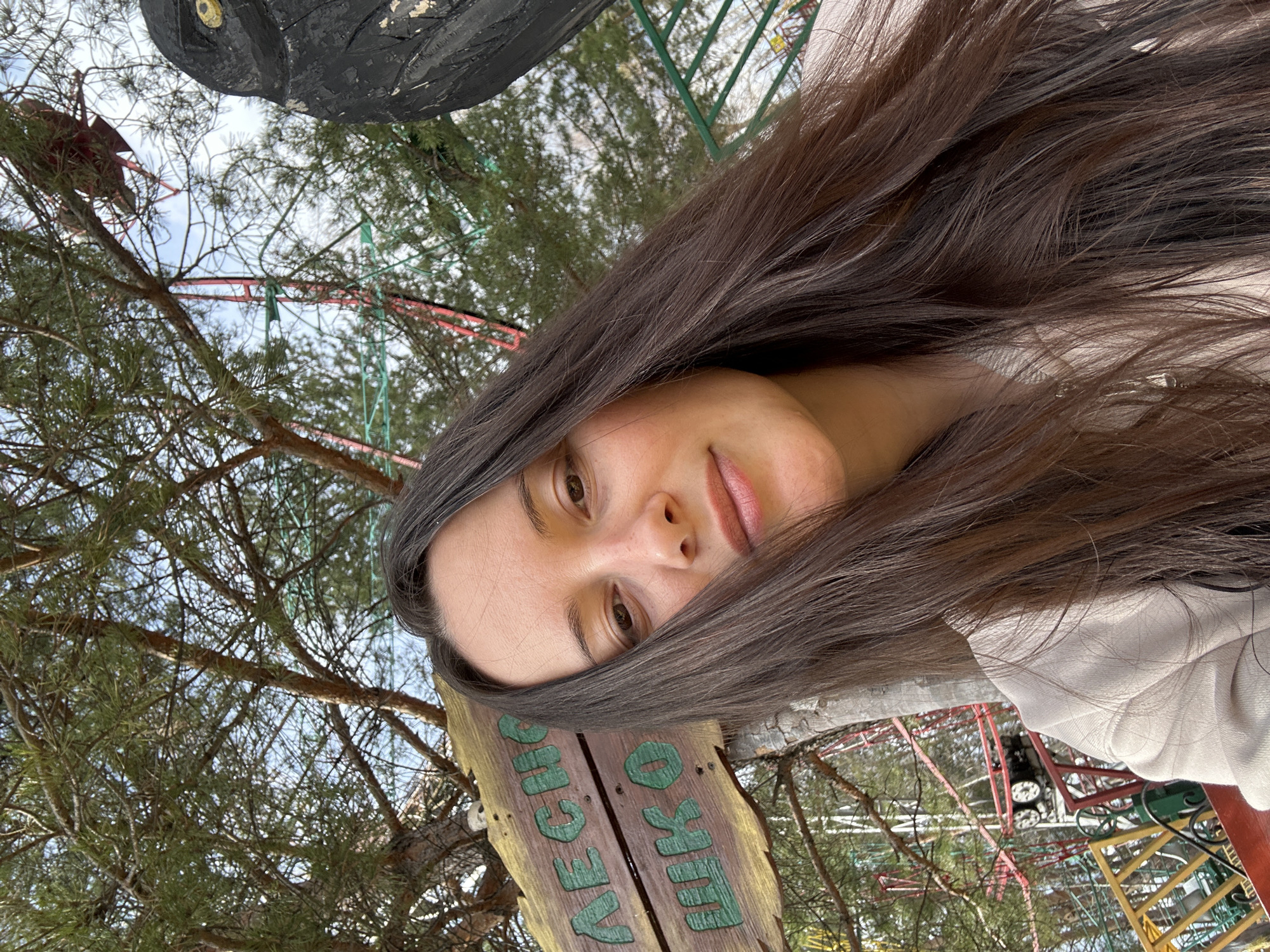 The image is a selfie of a woman with long, brown hair. She is outdoors, possibly in a park or amusement area, as indicated by the trees, amusement rides in the background, and a wooden sign written in Russian (likely directing to "Lesnaya Skazka"). The scene appears to be in Chaikovsky, Russia, based on this information. The subject is partially obscured by her hair and the sign.