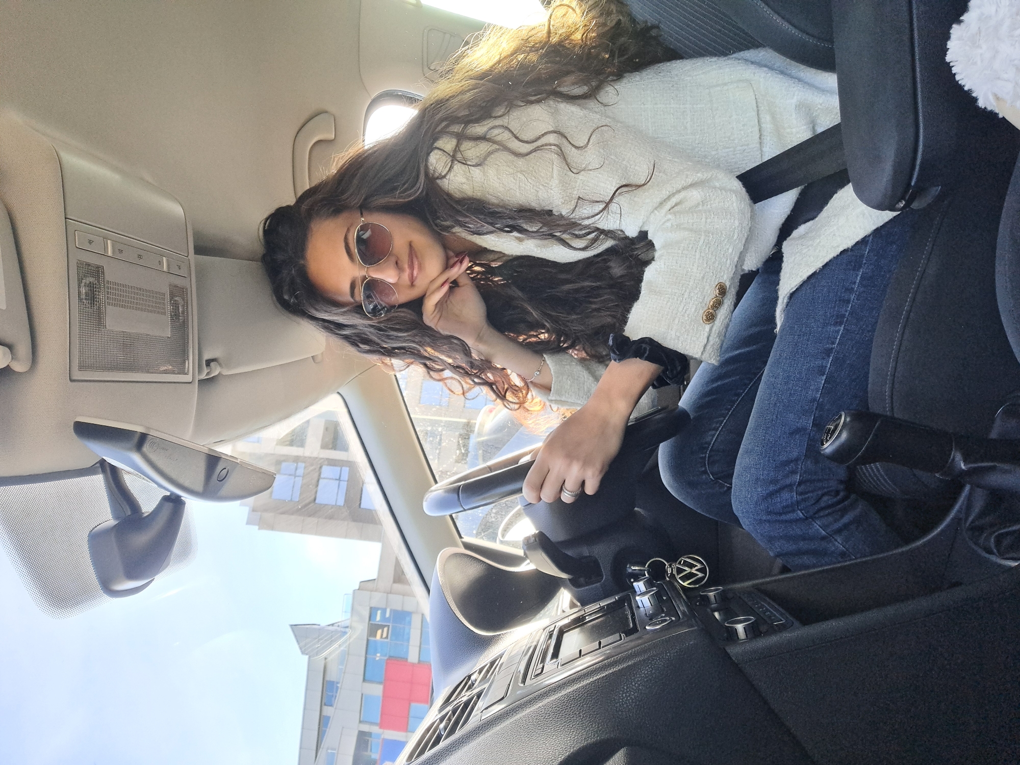 A woman is sitting in the driver's seat of a Volkswagen car. She is wearing a white tweed jacket, jeans, and sunglasses. She is holding the steering wheel with her left hand, which has a ring on its finger. Her right hand is near her face. The car is stopped in Sofia, Bulgaria, based on the architecture visible through the windshield.