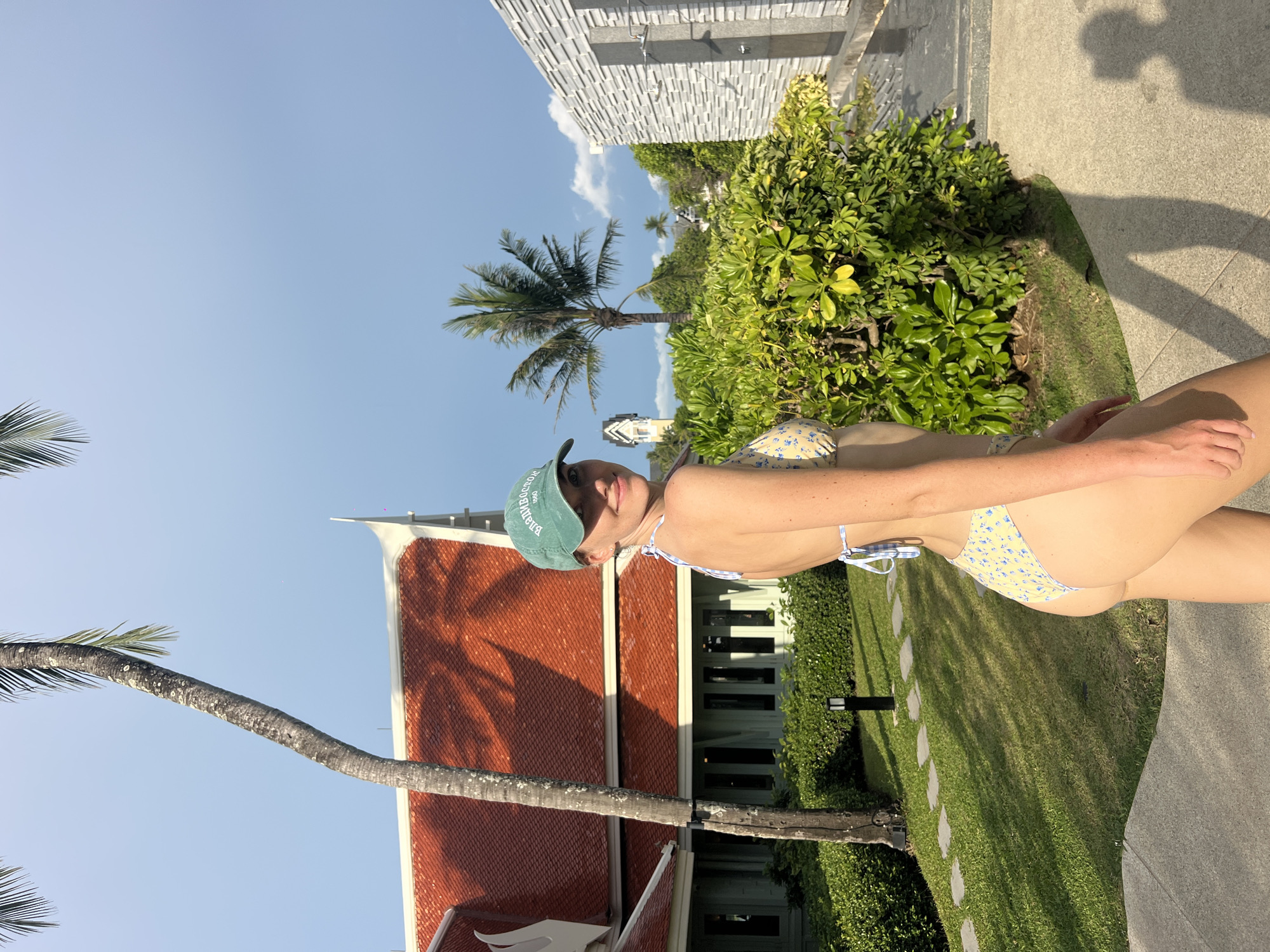 The image shows a woman in a bikini standing outdoors, likely in Choeng Thale, Thailand, given the Thai architectural elements visible. She is wearing a green baseball cap. Behind her is a building with a traditional Thai-style roof with orange tiles. A palm tree trunk extends across the foreground. The background includes greenery, a building with gray walls, and a blue sky with some clouds. The woman appears to be standing on a stone or concrete surface. A lawn and bushes border the scene.