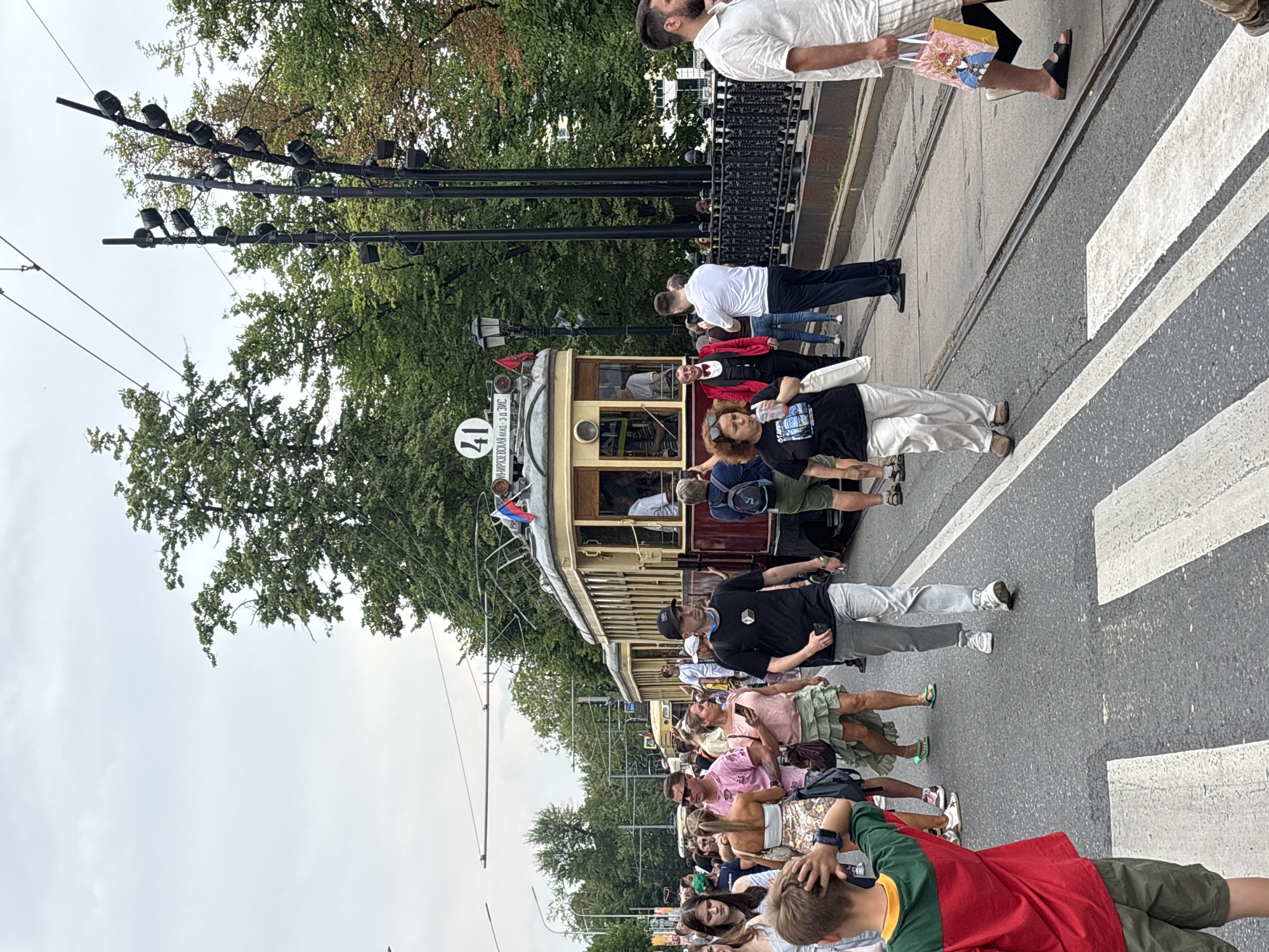 The image depicts a street scene in Moscow, Russia, likely during a public event or festival. A vintage tram, marked with the number 41 and the destination "Мир-Ульяновская" (Mir-Ulyanovskaya), is the central focus. The tram appears to be a historical exhibit or part of a parade, as suggested by the crowd and the festive atmosphere.
Numerous people are present, walking on the street, which has tram tracks running through it. Many are casually dressed, suggesting a warm weather day. Some individuals are looking towards the tram, while others are engaged in conversation or walking past. A young boy in a red, green, and yellow shirt is visible in the foreground, looking back and scratching his head. A man in a black t-shirt and grey pants is walking towards the viewer, and a woman in a black t-shirt and white pants is also walking in the same direction. Further back, a man in a red jacket with a bow tie is standing near the tram entrance, possibly an attendant or performer.
The environment includes trees lining the street, some with lush green foliage, and a cloudy sky. Streetlights with multiple bulbs are visible, and a decorative black metal fence runs along the side of the street. The presence of Russian flags on the tram indicates the location. The scene is bustling with activity, suggesting a lively public gathering.
