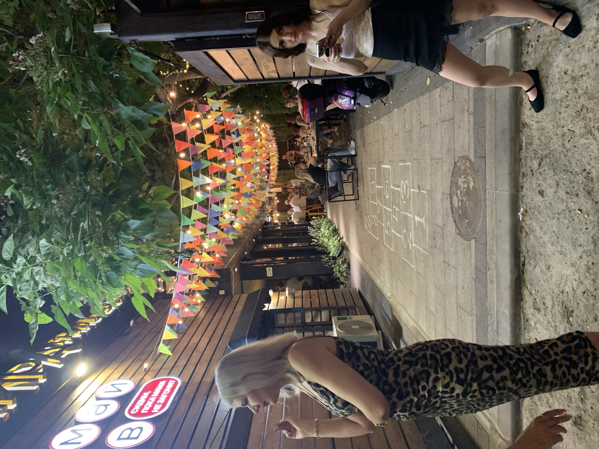 In the city of Unknown, Unknown, an outdoor dining area is depicted at dusk, illuminated by string lights and colorful pennant banners overhead. Two women are prominently featured in the foreground. The woman on the left, with blonde hair, wears a leopard-print dress and stands with her left hand near her mouth, appearing to hold a cigarette. The woman on the right, with dark hair, is dressed in a white top with a floral pattern and a black skirt with lace trim. She is looking at her smartphone, holding it in her hands. Behind them, patrons are seated at tables, with a purple scooter parked nearby. The ground is paved with cobblestones and features a hopscotch grid drawn in white chalk. The scene is set against a building with a wooden facade.