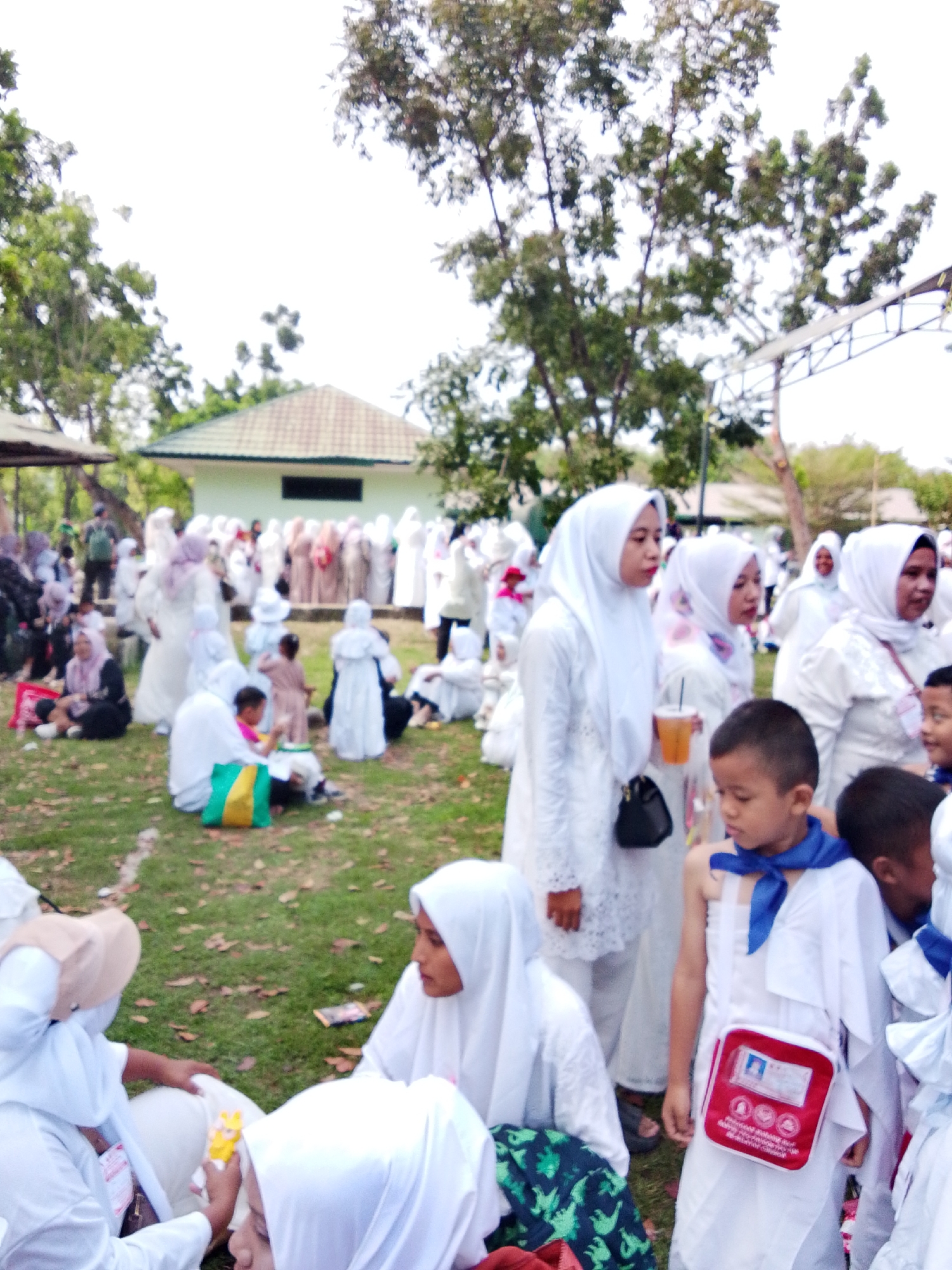 The image depicts a gathering of people, predominantly women and children, dressed in white attire, likely for a religious event, outdoors on a grassy area. Several individuals are seated on the ground, while others are standing. Some people are holding drinks, and one child is wearing a blue scarf around his neck and a red pouch on a strap. In the background, there is a building with a green roof and trees. The overall scene suggests a communal event taking place in a semi-rural or park-like setting. The specific location in Cirebon, Indonesia, is not visually discernible from the image itself, as there are no identifiable landmarks or signage.