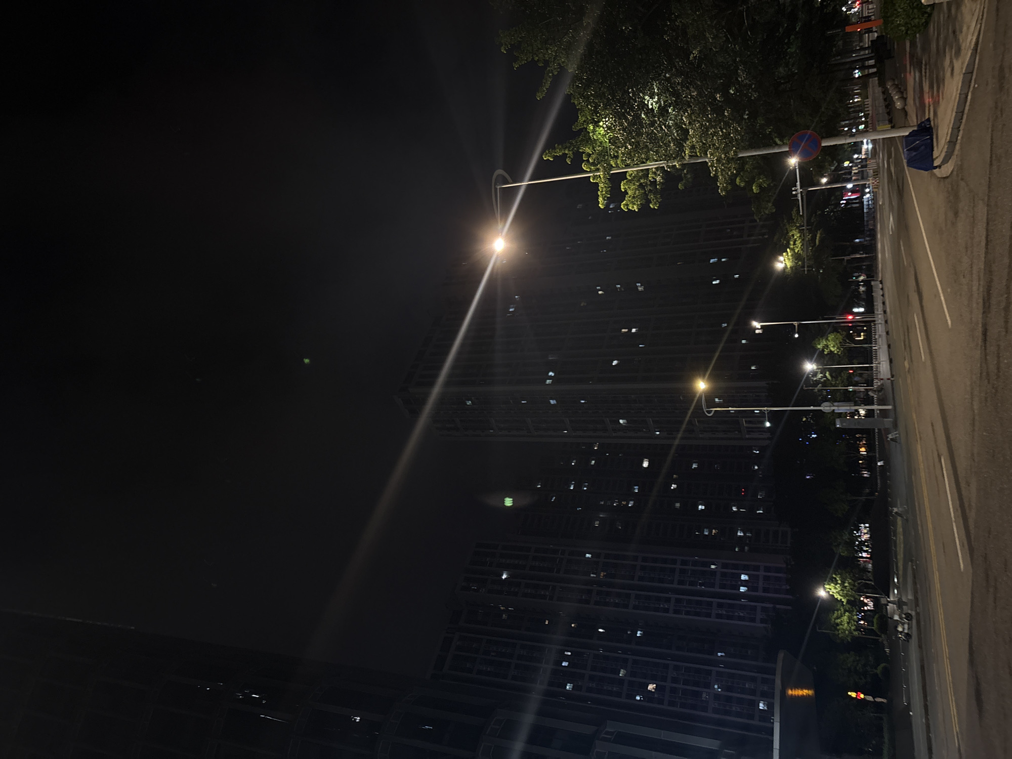 This media file captures a nighttime scene in Haizhu District, China, focusing on a city street bordering residential buildings and a park. The image is oriented vertically, with the street and buildings occupying the right and lower portions, while the sky and foliage dominate the left.
The primary subjects are the illuminated streetlights casting long streaks of light across the dark sky and reflecting on the wet pavement, suggesting recent rain. Several tall, modern apartment buildings with many lit windows are visible, indicating a populated urban environment. The architecture suggests a high-density residential area. A street lined with trees and lampposts separates the road from the buildings. In the foreground of the street, a blue trash bag is placed next to a lamppost. A circular traffic sign with red and blue markings is also visible on a lamppost.
The time of day is clearly night, with the darkness emphasized by the artificial lights. The wet road and the lush greenery of the trees suggest a humid climate, possibly during or after a period of rain. The scene is peaceful and quiet, with no visible people or activity, evoking a sense of urban stillness after dark. The city lights create a somewhat surreal atmosphere. No legible text is present in the image.