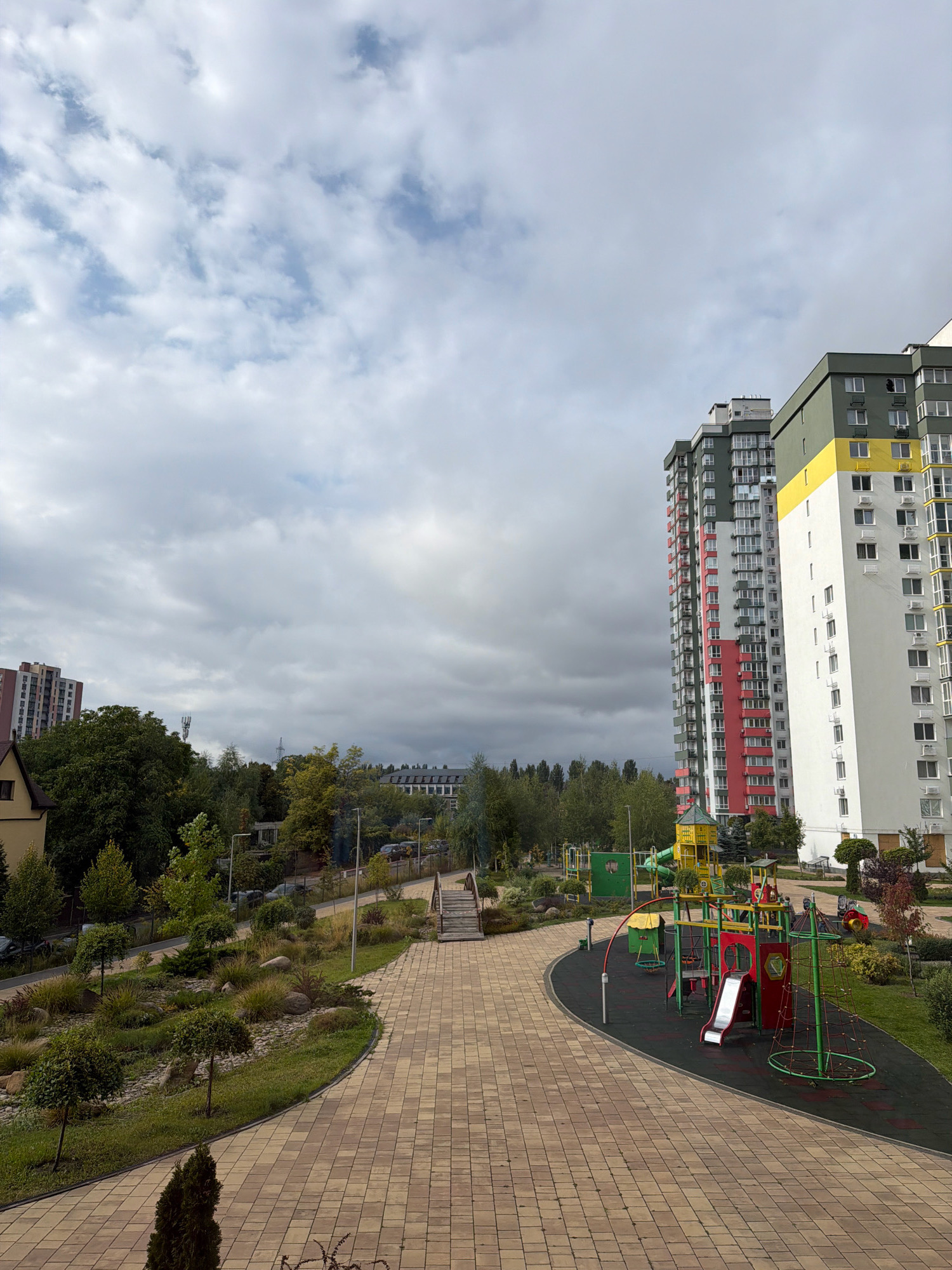 The image depicts a modern residential area with a focus on a well-maintained playground and surrounding landscaping. In the foreground, a paved pathway made of tan square tiles curves through a landscaped area with various plants, small trees, and rocks. To the right of the pathway is a colorful children's playground with slides, swings, and climbing structures, set on a black rubberized surface.
Dominating the background are several tall, modern apartment buildings. The building closest to the viewer is white with a yellow band near the top and a green band below that. To its left is a taller building with sections of red and green facades, and further left, another apartment building with a patterned red and white exterior.
The environment appears to be an urban or suburban setting, with a cloudy sky overhead. Trees and vegetation are visible throughout the scene, suggesting a green and residential development. The overall impression is one of a contemporary, community-oriented living space.
