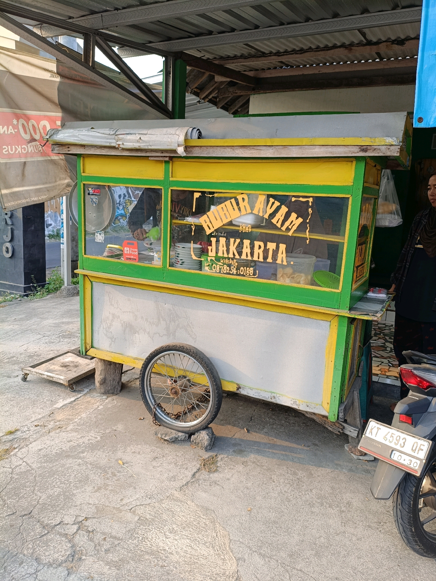 The image depicts a food cart for "Bubur Ayam Khas Jakarta" (Chicken Porridge typical of Jakarta) parked on a concrete surface in Maguwoharjo, Indonesia. The cart is painted in bright yellow and green, with glass windows displaying food items and utensils. A person wearing a dark hijab and a plaid shirt stands to the right of the cart, near a motorcycle with a license plate reading "KT 4593 QF". The cart has a single wheel on the right side and a small platform with wheels on the left. In the background, a corrugated metal roof shelters the cart, and a banner with red lettering and the number "000" is visible on the left. The environment appears to be an outdoor setting, possibly a street or a marketplace, with a paved ground and some vegetation. The lighting suggests it is daytime, possibly late afternoon due to the warm tones.