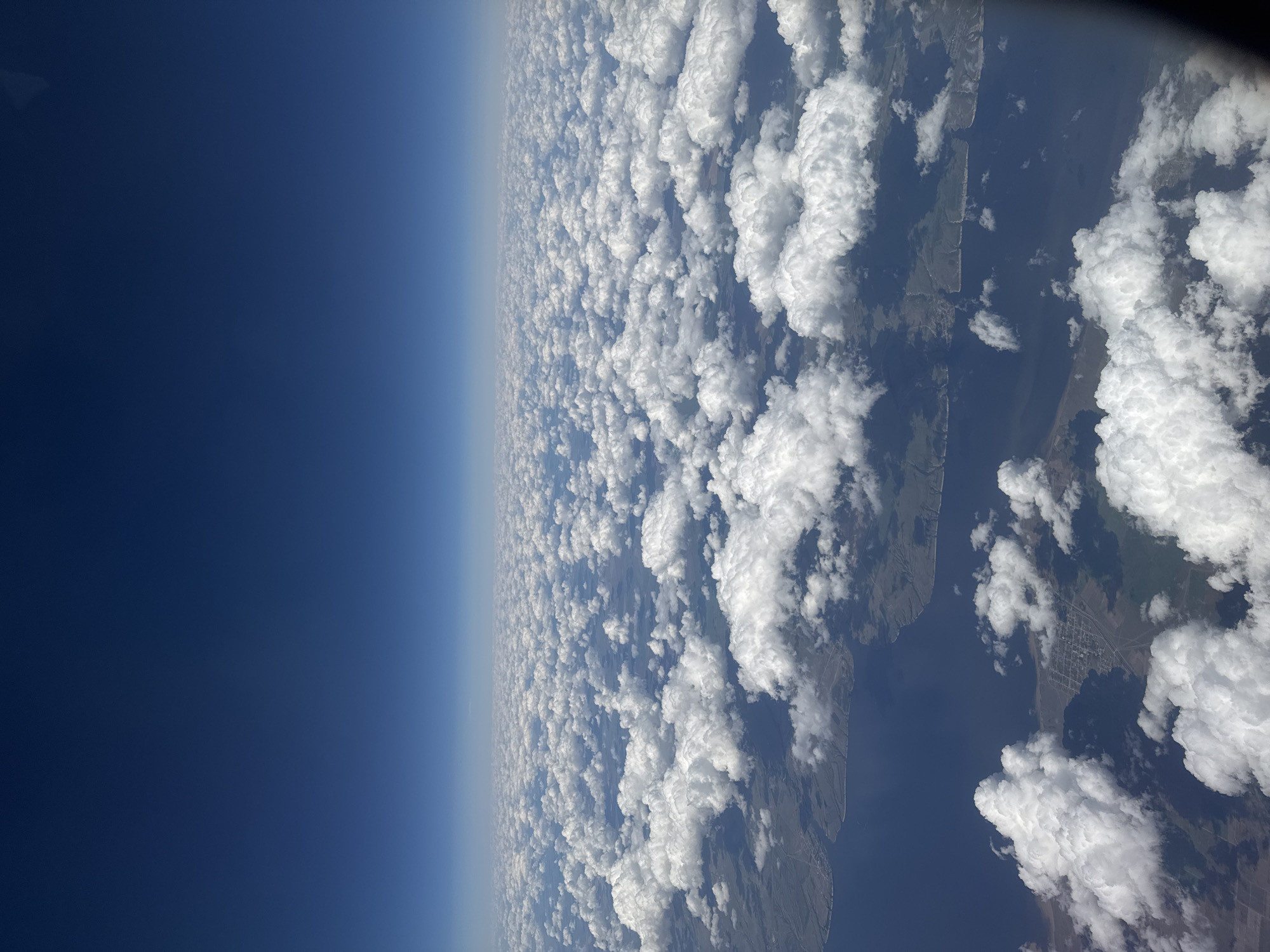 The image shows a view of Earth from a high altitude, possibly from an airplane or spacecraft. The left side of the image is dominated by a dark blue expanse, representing the upper atmosphere or space. A gradient transitions from this dark blue to a lighter blue, indicating the edge of the atmosphere. The right side of the image features a dense layer of white clouds, obscuring much of the ground below. There are, however, what appears to be some bodies of water and land visible through gaps in the clouds. It is not possible to identify the city of Izhevsk, Russia, without ground-level imagery.