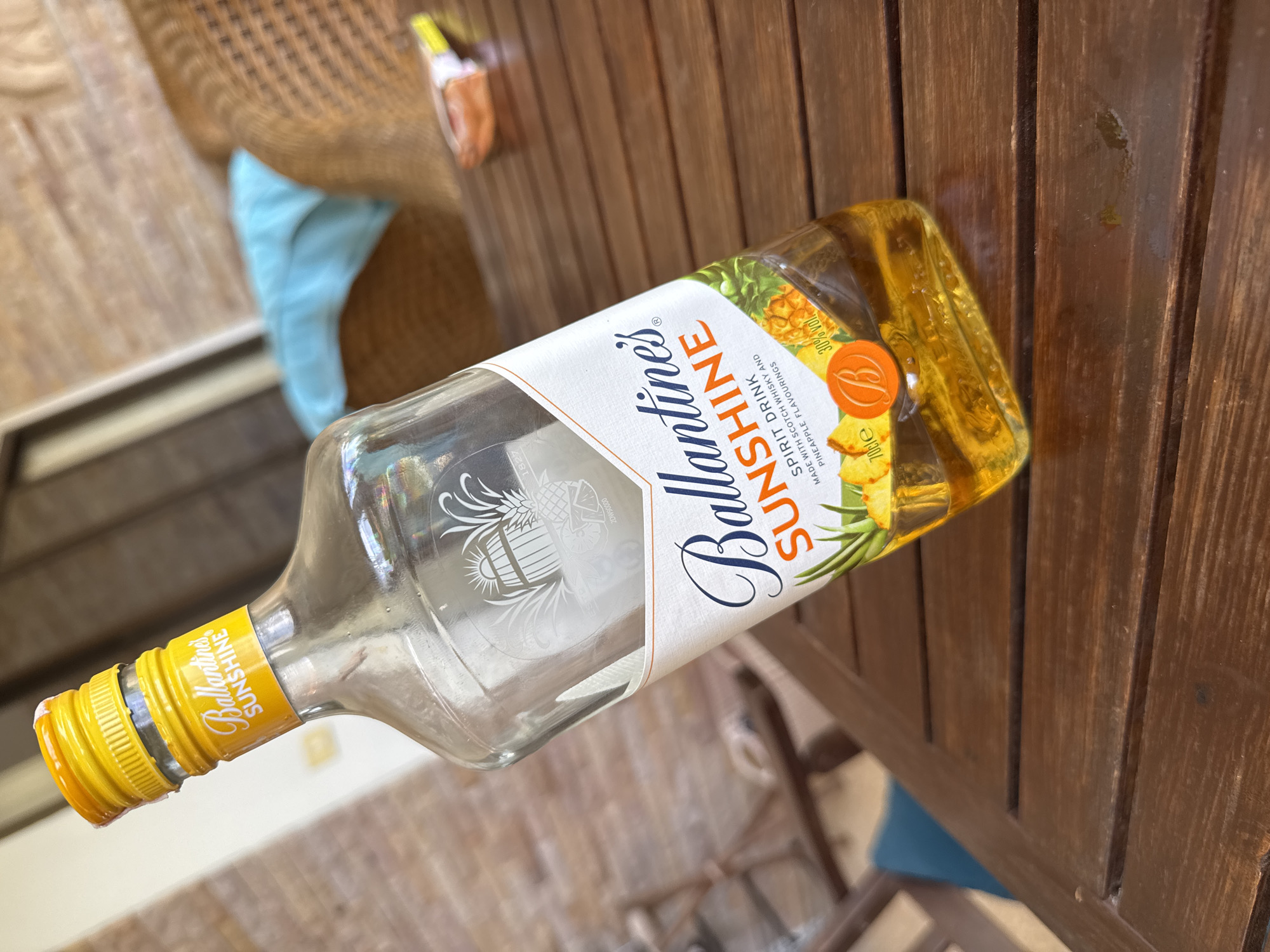 A tilted bottle of Ballantine's Sunshine spirit drink sits on a brown wooden table. The bottle is mostly empty, with a yellow cap and a label featuring pineapple imagery. The table is composed of vertical wooden slats. In the background, parts of a chair with woven material and a blue cushion are visible. The image was likely taken outdoors, and while the location is near Rawai, Thailand, this cannot be verified by the image.