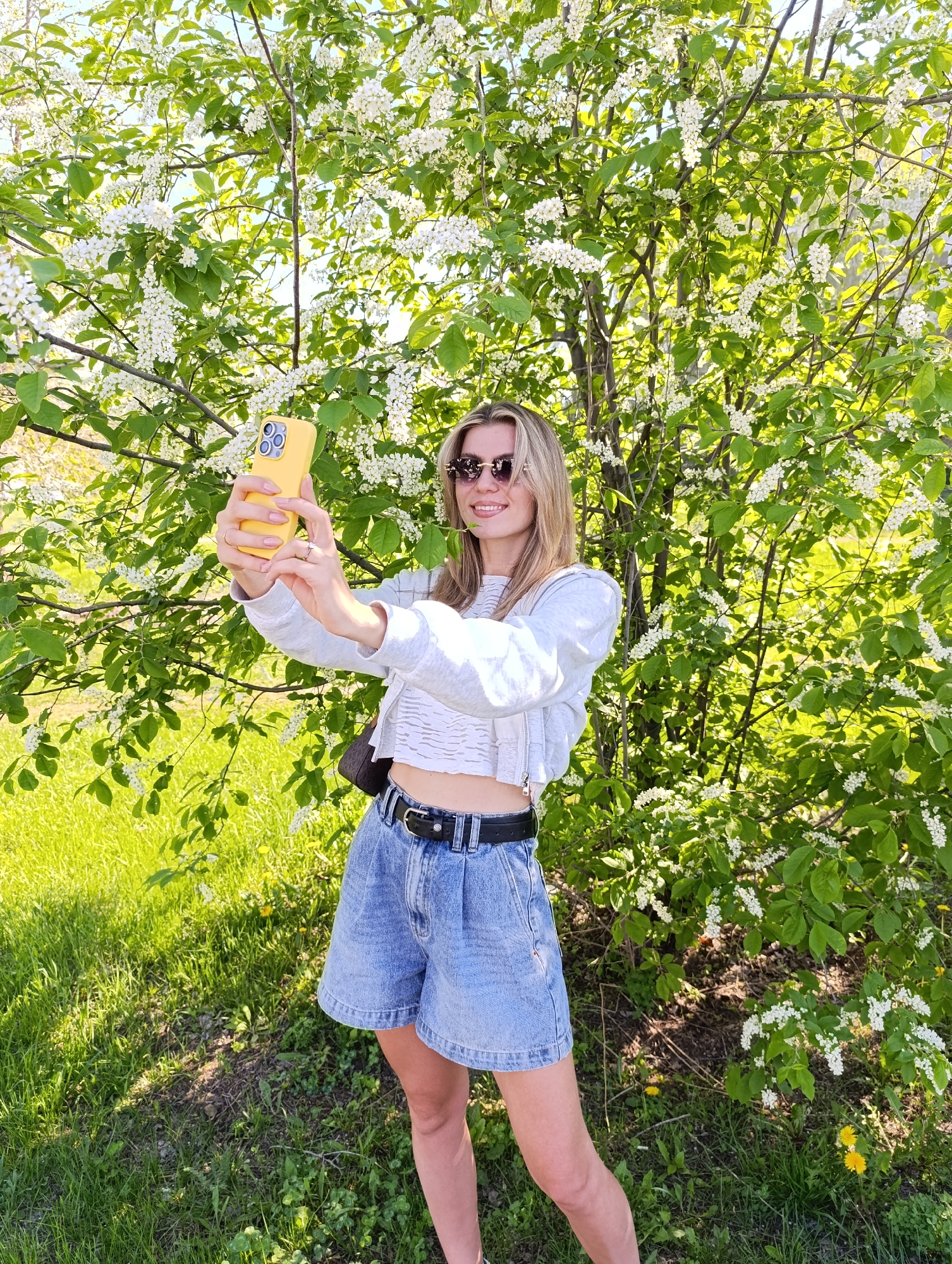 A woman stands in a grassy area with a tree covered in white flowers behind her. She is fair-skinned with light blonde hair, wearing sunglasses, a cropped white shirt under a light grey zip-up sweater, denim shorts with a black belt, and a dark shoulder bag. She holds a yellow phone up, presumably taking a photo or video of herself. The location is a park or field, and the sky is not visible but appears to be a sunny day.