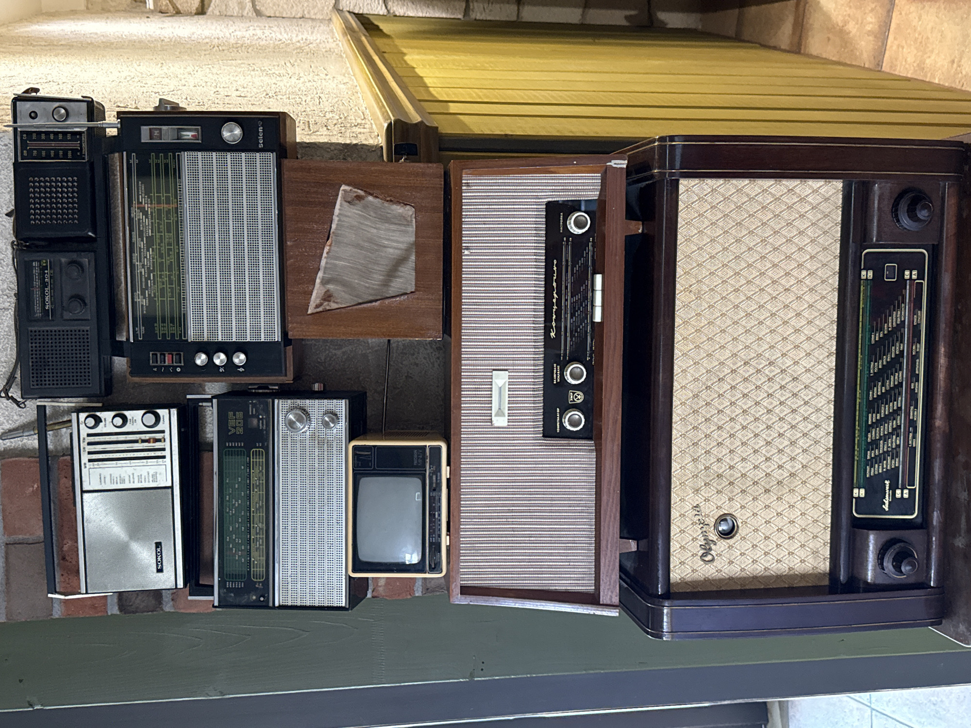 The image shows a stack of vintage radios and a small television against a brick wall. The objects are arranged vertically, with the smallest radio at the top and the largest radio at the bottom. The radios vary in size, color, and design, with some featuring metallic fronts and others wooden casings. The small television is yellow and appears to be from the mid-20th century. The background is a brick wall, and to the right, there is a section of yellow-painted wooden paneling and a beige stone wall. The lighting is somewhat dim, suggesting an indoor setting. The scene is static and does not depict any people or ongoing actions. The overall impression is a display of retro electronic devices, possibly in a vintage shop or a private collection. The location in the city of Pleven, Bulgaria is not evident from the visual information provided in the image.