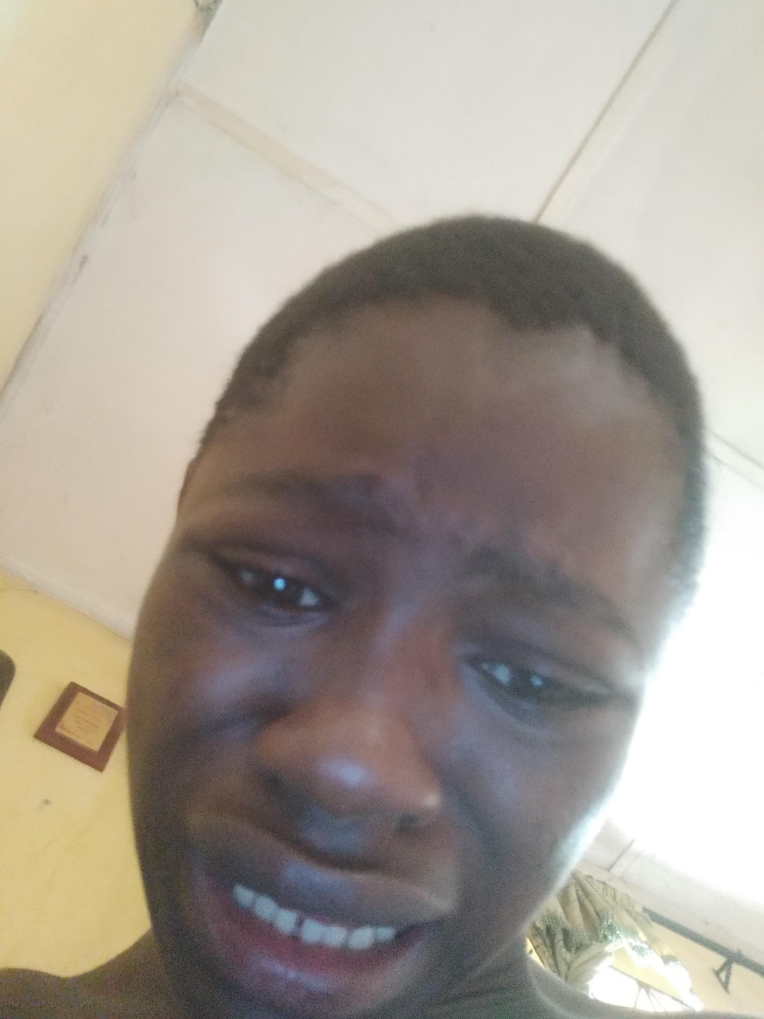 A close-up selfie of a young Black person with short, dark hair. The person's face shows a strained or upset expression, with furrowed brows and clenched teeth. The background is out of focus, showing parts of a white ceiling and a yellowish wall with a framed object. The location, Shangisha, Nigeria, is provided but is not directly discernible from the visual elements in the image.