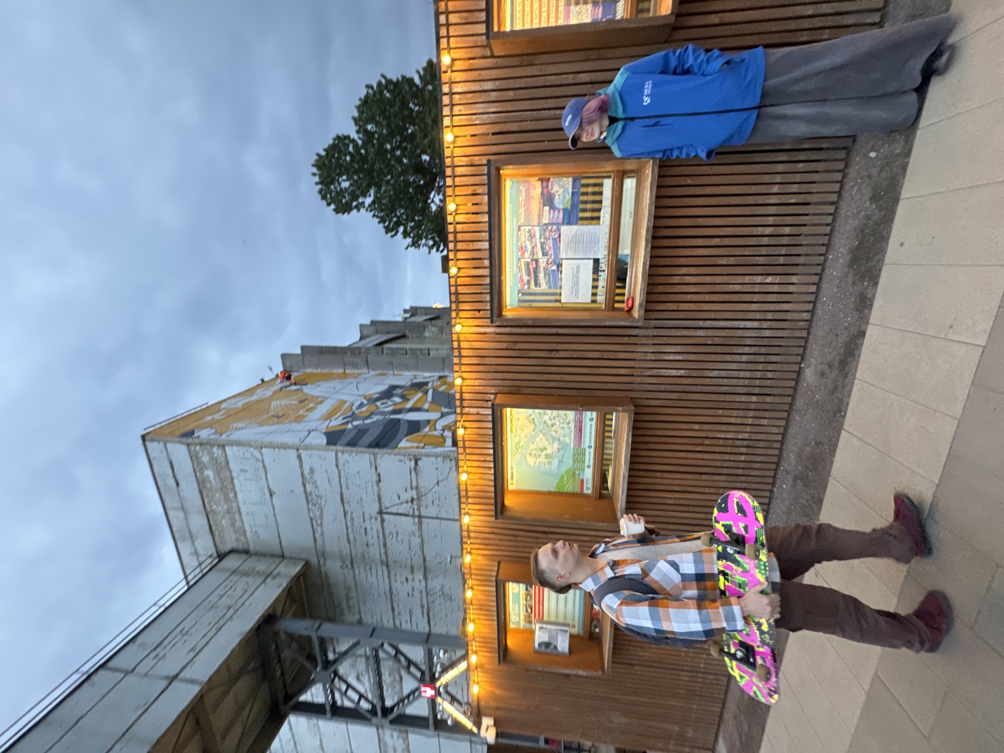 Two people are standing in front of a wooden wall with illuminated display cases. The person on the left is a young man wearing a plaid shirt, brown pants, and dark shoes, carrying a skateboard and a cup. He is looking to the right. The person on the right is wearing a blue jacket, a purple hat, and a long grey skirt. They are looking to the left. The wooden wall has vertical slats and is decorated with string lights. The display cases contain posters or information. Behind the wall, there is a building with large windows and artwork on its exterior. A pedestrian walkway is visible in the foreground. The sky is overcast. The image appears to be taken in Saint Petersburg, Russia, based on the context provided.