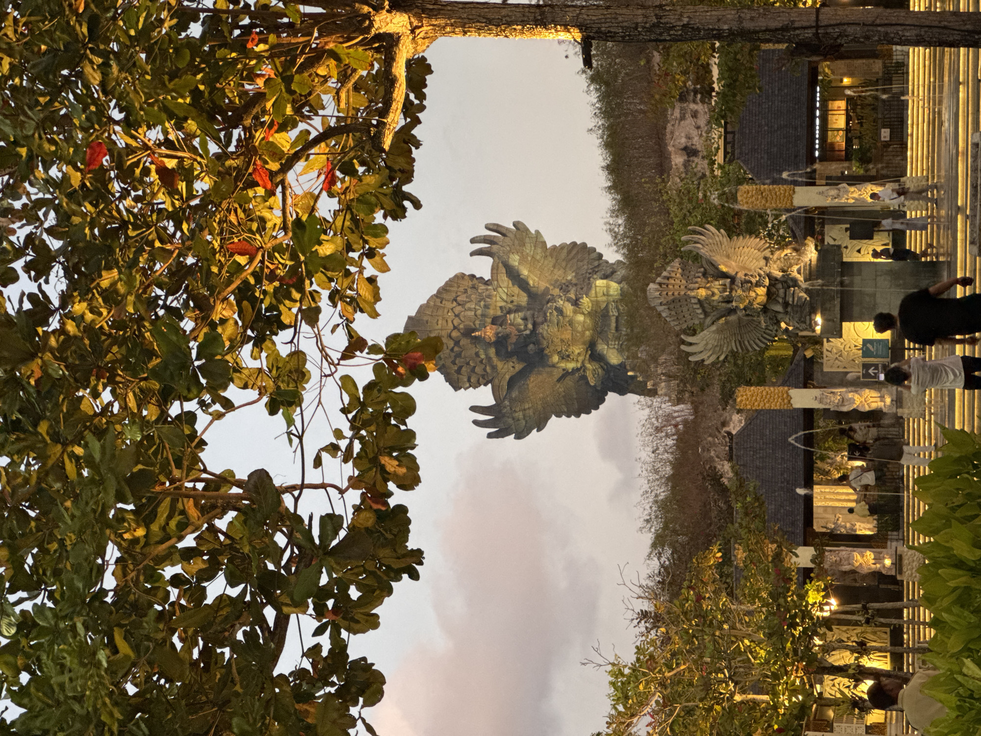 The image shows the Garuda Wisnu Kencana (GWK) Cultural Park in Kuta Selatan, Indonesia, at dusk. In the background, a colossal statue of Garuda, a mythical bird-like creature, is illuminated. In the foreground, there are several smaller statues, fountains, and people. The area is surrounded by trees and foliage. The sky is overcast with muted colors. People are seen walking around, taking photos, and observing the statues. The overall atmosphere is serene and culturally rich.