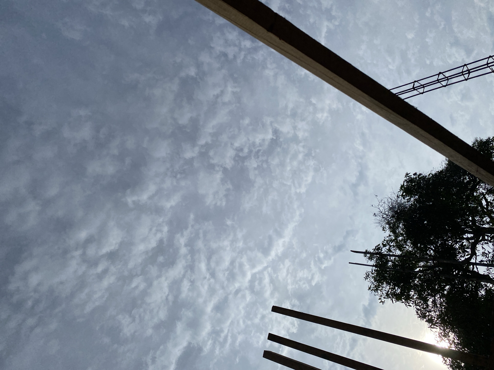 The image shows a view looking up at a cloudy sky. In the foreground, there are several construction elements. On the left, three dark vertical poles are visible. A diagonal wooden beam extends from the right side of the frame towards the center, with reinforcing bars attached to its underside. In the background, silhouetted against the sky, is a large tree with dense foliage. The sun is peeking through the clouds on the left side of the image, casting a lens flare.