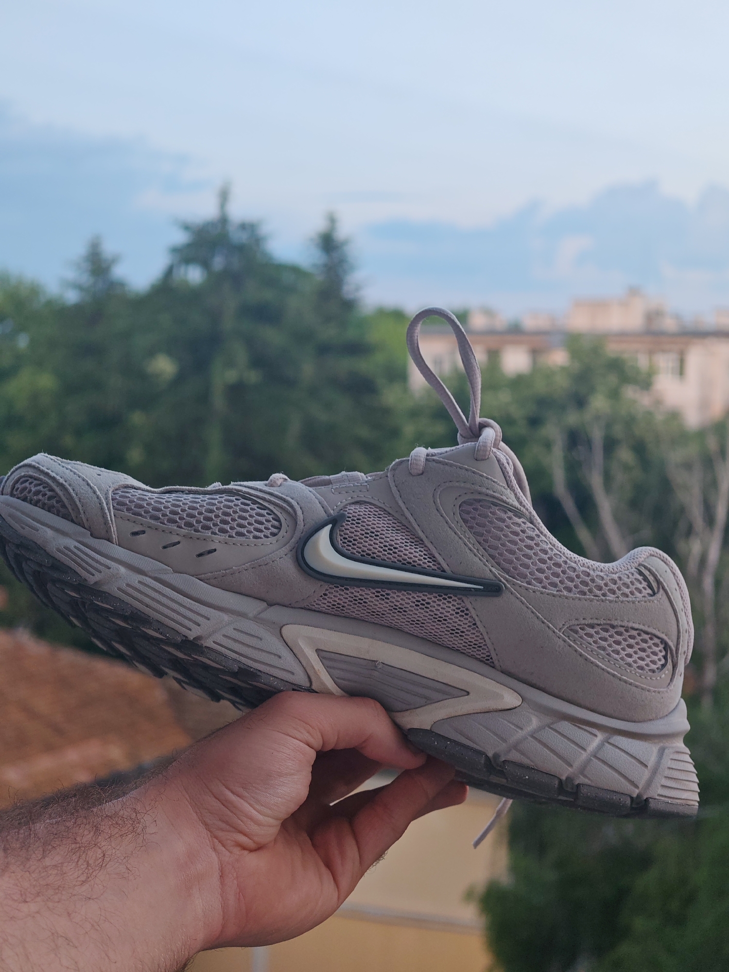 A male individual's right hand, visible with arm hair, holds a single light grey/taupe Nike athletic shoe, featuring mesh and suede-like overlays, against an out-of-focus outdoor background. The shoe's white-filled Nike swoosh logo, outlined in black, is prominent on its lateral side, and the laces are looped at the top. The sole exhibits visible wear and dirt. The background consists of a light blue sky with diffused clouds in the upper portion, dense green foliage (trees) in the mid-ground, and light-colored urban buildings with multiple windows partially visible through and above the trees on the right side. A blurred reddish-brown surface, possibly a terracotta roof, is seen in the lower left beneath the hand and shoe. The scene is located in the city of Shumen, Bulgaria.