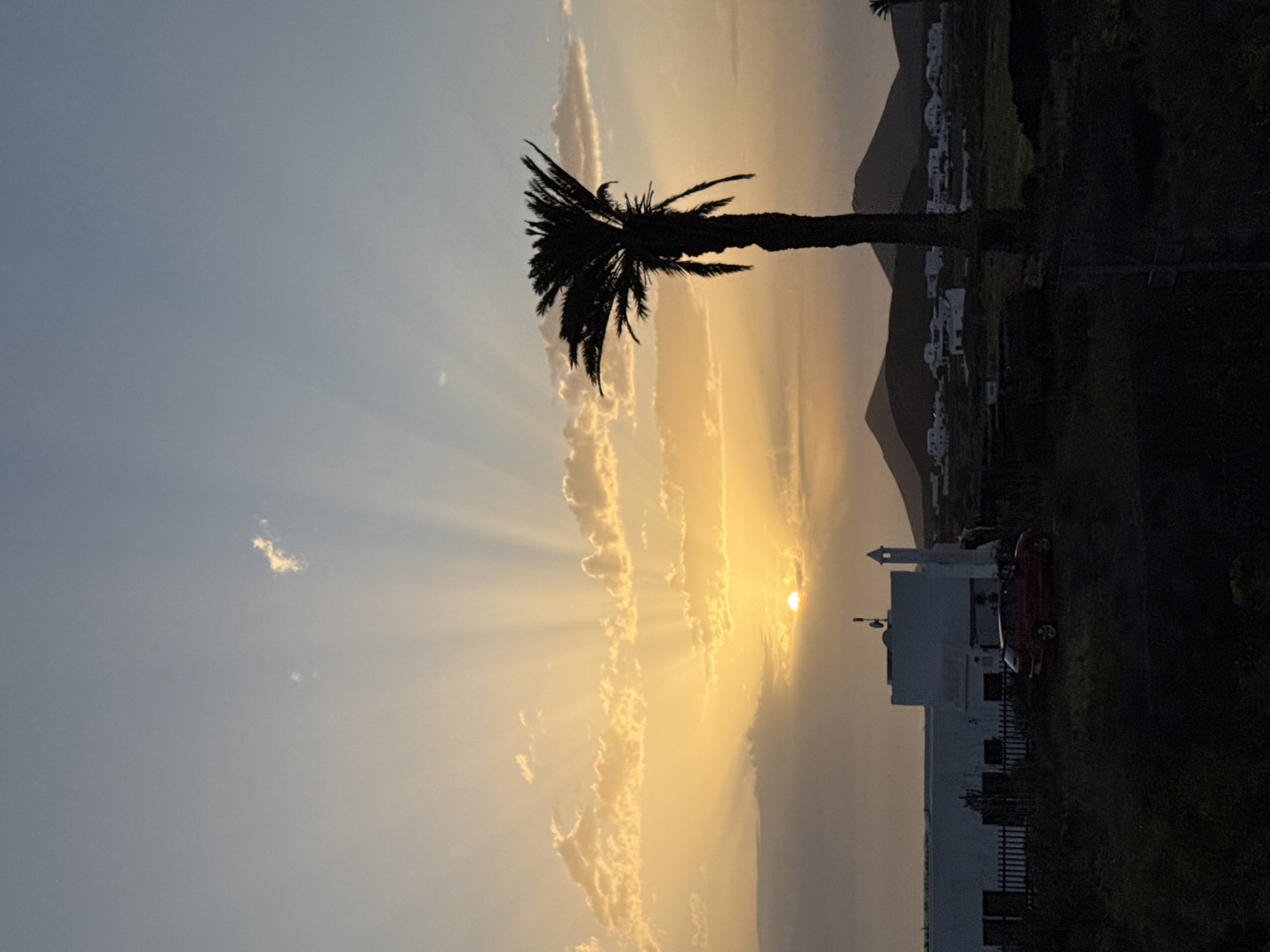 The image is a landscape photograph taken in Yaiza, Spain, depicting a sunset. A tall palm tree is silhouetted against the golden light. Behind the tree, there's a building with a tower, also silhouetted, along with a red car. In the distance, hills and small white houses are visible. The sky has a blue gradient, with light rays emanating from the setting sun, obscured by clouds.