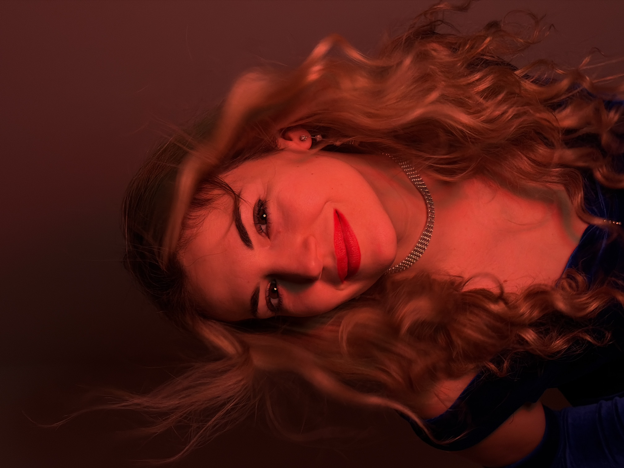 A low-angle shot depicts a fair-skinned woman with long, wavy blonde hair and bright red lipstick. She wears a silver chain necklace and a dark blue velvet top. Her head is tilted towards the viewer and slightly to the right, with her left eye in focus. The lighting is predominantly red, creating a warm, saturated tone on her skin and hair. The background is dark and out of focus. There is no indication of location in Budva, Montenegro. The image is a portrait.