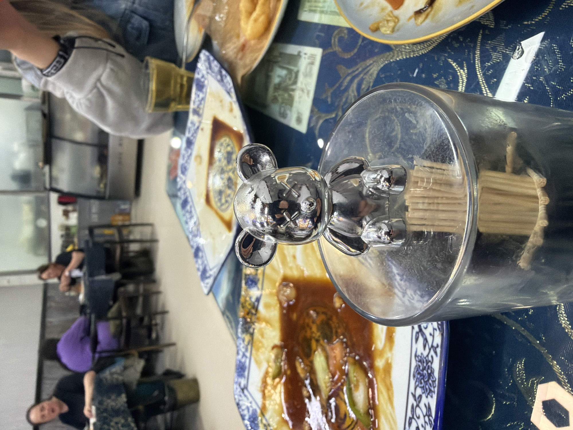 The image, oriented vertically, depicts a lively indoor dining scene, likely within a restaurant or home in Tongjiang, China. The focal point is a reflective, chrome-plated bear-shaped figurine, reminiscent of a Be@rbrick or KAWS Companion with distinct "X" marks for eyes, perched on top of a cylindrical glass container filled with wooden toothpicks.
The dining table is adorned with a dark blue tablecloth featuring intricate gold patterns. Several ceramic plates with traditional blue and white Chinese designs are present. One plate directly beneath the toothpick holder holds remnants of a meal, including a rich brown sauce with sliced green onions and other ingredients, suggesting a savory dish. Another plate further back also shows signs of having held food.
In the mid-ground, a blurred green banknote, likely Chinese Yuan given the location context, is visible on the tablecloth beneath a plate, displaying indistinct characters and numbers. A glass cup containing a light amber liquid is also on the table.
The background is softly blurred but reveals several people and parts of the room. One individual, wearing a grey long-sleeved top and blue jeans, is seated nearby, with a black and white patterned wristband visible on their arm. Further back, two other people are vaguely discernable: one in a black top and purple pants, and another in a black top. The setting suggests a casual meal, with dishes partially consumed under artificial indoor lighting.