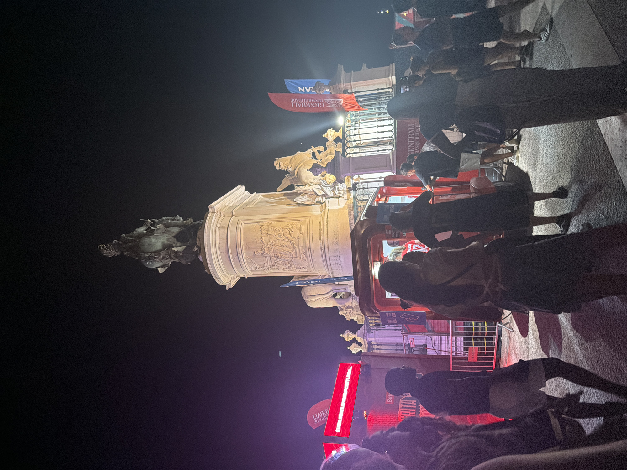 The image depicts a nighttime scene in Lisbon, Portugal, likely at Praça do Comércio, a prominent public square. In the background, a large, ornate monument featuring a statue of a man on horseback dominates the skyline. The monument is illuminated and surrounded by architectural elements. In the foreground, a crowd of people are gathered, suggesting an event or public gathering. Red barriers are visible, indicating a designated area or queue. Several large red banners with white text are displayed, one of which reads "SUMMA GESTÃO DEPORTIVA PORTUGAL". Flags of Generali, an insurance company, are also prominently displayed, one red and one blue. A red booth or structure, possibly a temporary stage or information point, is present. The overall atmosphere appears festive, with artificial lighting creating a dramatic effect against the dark night sky.