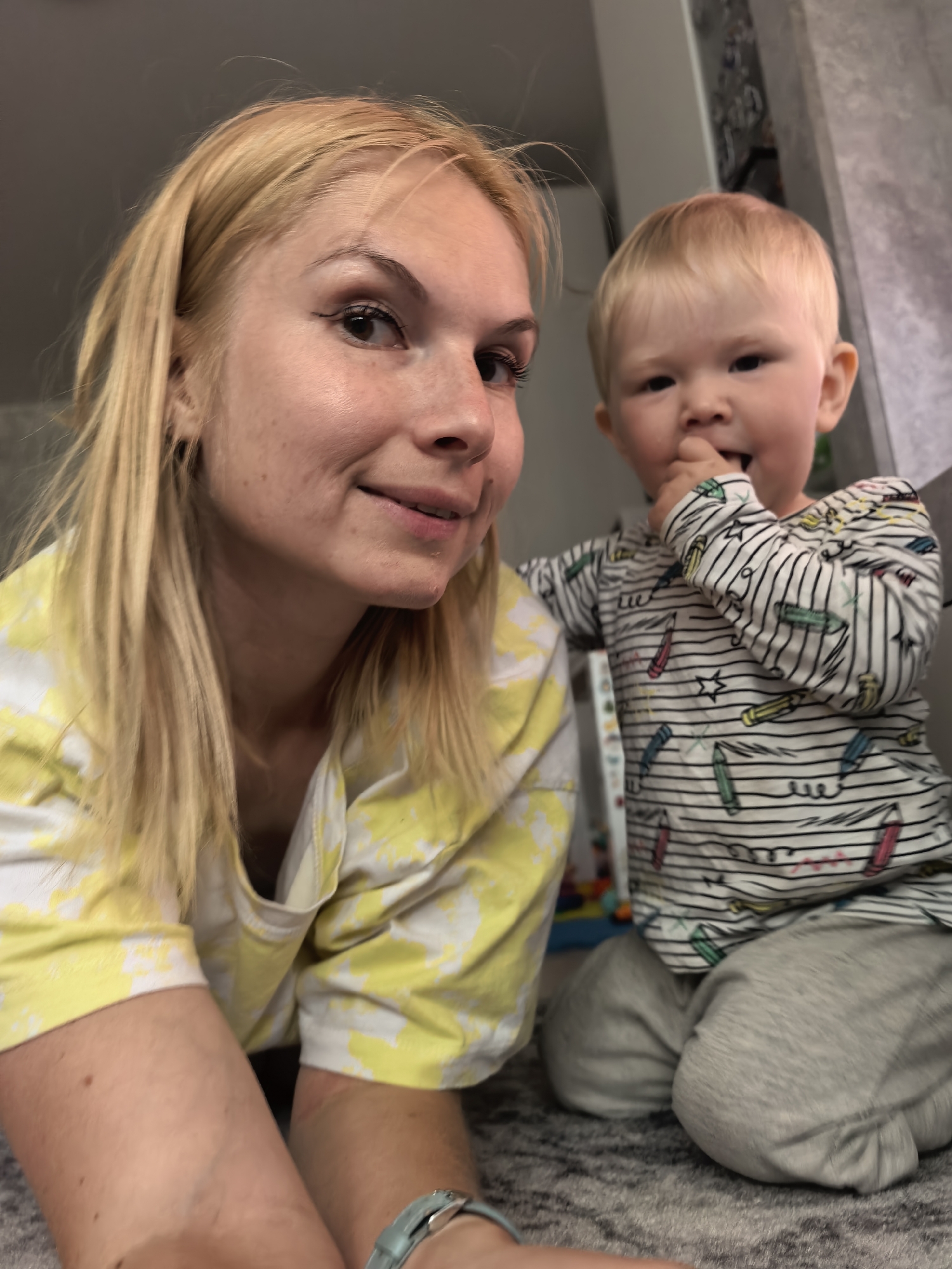 A woman with blonde hair and a toddler are kneeling on a carpeted floor indoors. The woman is on the left side of the frame, facing forward with a slight smile. She is wearing a yellow and white tie-dye shirt and a watch on her left wrist. The toddler is on the right, wearing a striped long-sleeved shirt with crayon designs and gray pants. He is holding his hand to his mouth. The background is out of focus and appears to be a wall or furniture. The location is Sterlitamak, Russia.
