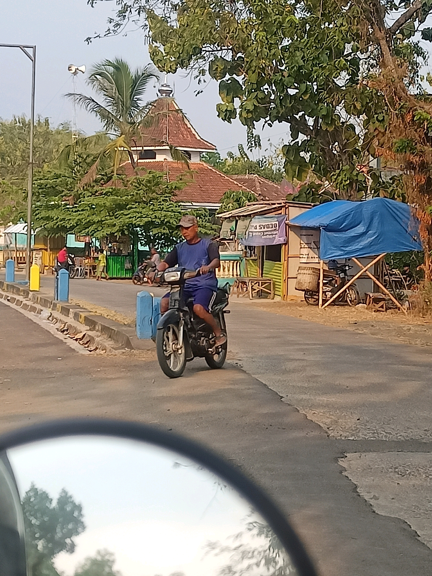 A man wearing a baseball cap, blue t-shirt, and blue shorts rides a black motorcycle down a paved road. The motorcycle has a silver exhaust pipe and silver rims. In the background, there are buildings with red tile roofs, including what appears to be a mosque with a domed roof. Several trees with green leaves are present, along with a blue tarp stretched over a wooden frame. A white speaker is visible hanging from a pole on the left side of the image. The scene is set during daylight, with the sky appearing light blue. There are yellow and blue bollards lining the side of the road. A portion of a car's side mirror, reflecting trees and the sky, is visible in the foreground. The overall setting suggests a rural or semi-urban area in Sambiduwur, Indonesia, possibly near a local market or community center.