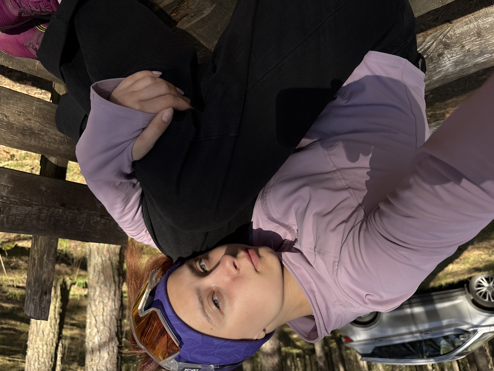 A woman is sitting on a wooden bench outdoors, possibly in a wooded area. She is wearing black pants, a light purple long-sleeved shirt, and pink and black shoes. She has goggles on her head. In the background, trees and a portion of a silver car are visible. The specific location in the city of Perm, Russia, is not discernible from the image.