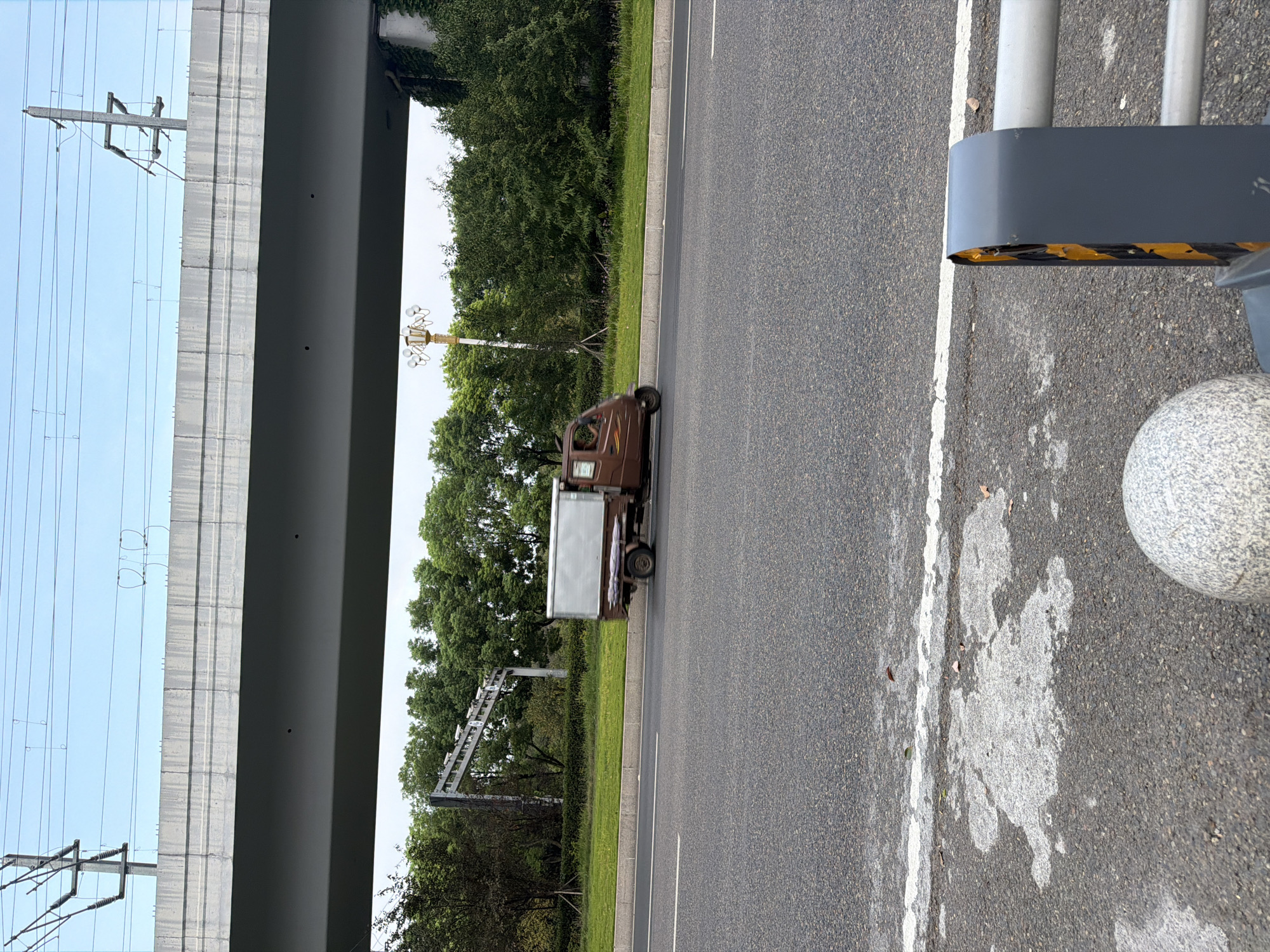 A brown three-wheeled truck with a silver cargo box is driving on a road in Jiangjin, China. The truck is in the middle of the frame, slightly to the left. Behind the truck, there is a line of trees and a large overpass. The overpass is made of concrete and has power lines running above it. In the foreground, there is a grey sphere and a section of a metal barrier. The sky is clear and blue.