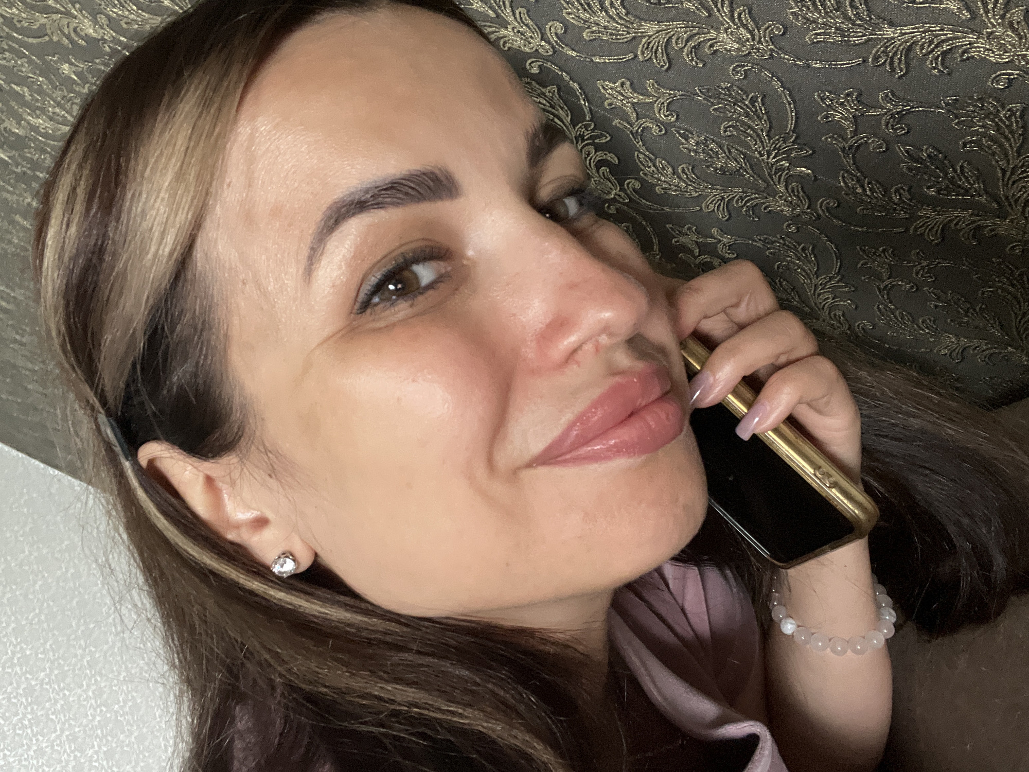 A woman is lying on her side, her face close to the camera. She has long, dark brown hair with lighter brown highlights, and she is wearing a pink shirt. On her left ear, she has a diamond stud earring. Her right hand is holding a gold-colored phone, and she is wearing a bracelet on her left wrist made of white, translucent beads. Her makeup includes defined eyebrows, eyeliner, and pink lipstick. She is resting her head on a textured, dark gray surface with a gold damask pattern. The overall lighting suggests an indoor setting.