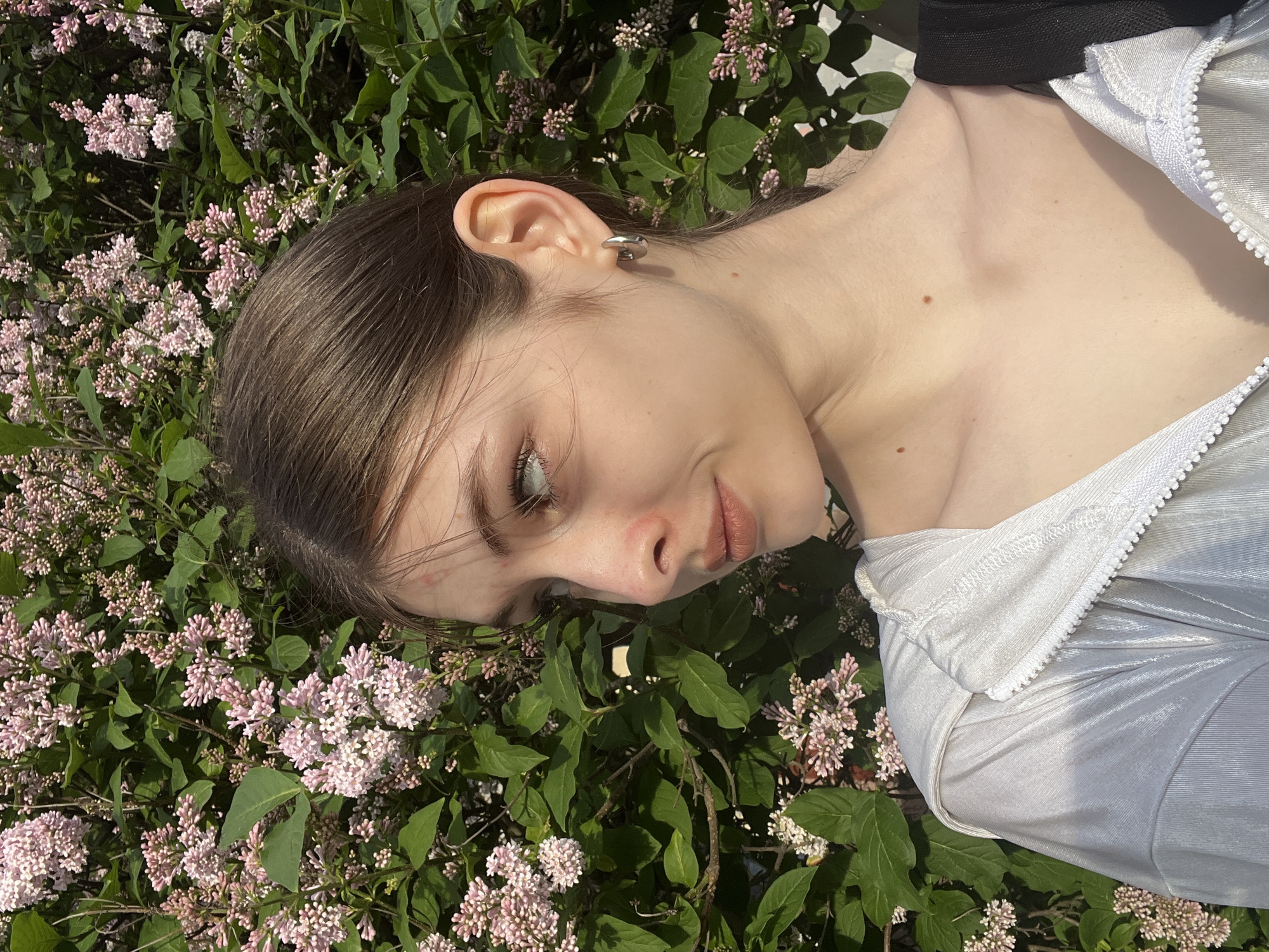 The image captures a close-up portrait of a young woman lying down amidst a dense growth of flowering plants. She is positioned on her side, with her head gently resting among lush green leaves and abundant clusters of small, pale pink to light lavender flowers, strongly resembling lilacs.
The woman has brown hair, styled back to reveal her ear, which is adorned with a distinctive silver-toned ear cuff or earring. Her face is presented in profile, looking upwards and slightly away from the camera, with a calm and soft expression. Her skin appears fair, and the natural daylight illuminates her features, casting subtle shadows and highlighting her contours.
She is wearing a light-colored, possibly silver or metallic light blue, garment with a prominent white zipper detail visible along its edge, suggesting a jacket or top. A black band or strap is visible near her shoulder and neck. Her exposed chest shows two small, distinct moles.
The setting is outdoors, under bright, natural sunlight, indicating either a morning or afternoon scene. The vibrant green foliage and delicate flowers create a serene and natural backdrop. The image conveys a sense of tranquility and natural beauty, possibly taken in a garden or park in Saint Petersburg, Russia, during the season when lilacs are in bloom. There is no visible text in the media file.