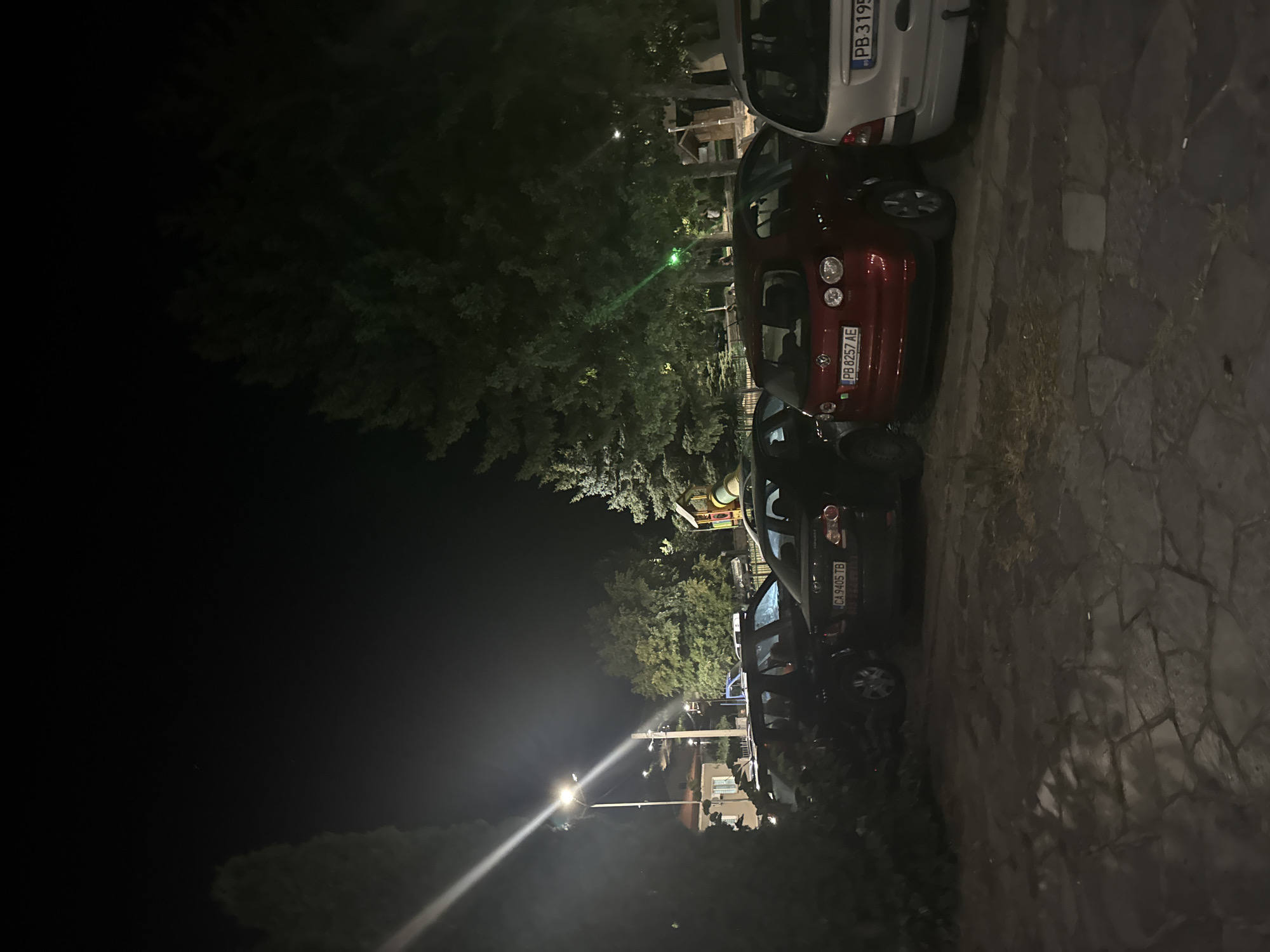 The image shows a nighttime scene in the city of Sopot, Bulgaria. In the foreground, a dark-colored SUV is parked. To its right, a red Volkswagen Golf is parked, with its license plate reading "PB 8257 AE". To the right of the Golf, a silver car is parked, with its license plate partially visible as "PB 3195". Behind these cars, a playground structure with colorful slides is visible amongst trees. A street light illuminates the scene, casting long shadows. The ground is paved with irregular stone slabs.
