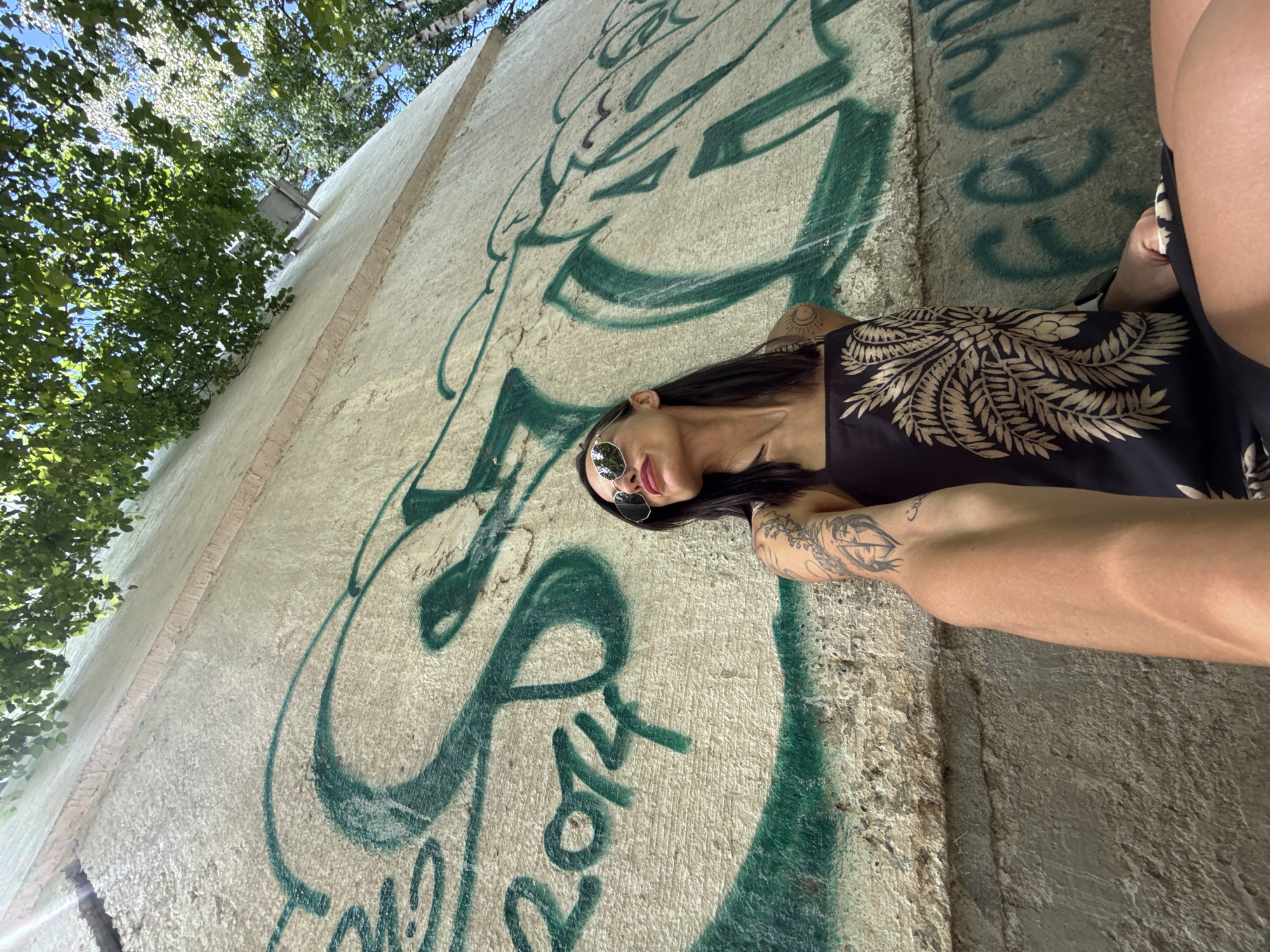An outdoor, low-angle shot captures a woman lying or leaning against a graffiti-covered wall, looking towards the camera. The image is rotated, showing the woman positioned along the wall, with her head towards what appears to be the bottom left of the frame.
The subject is a woman with long, dark hair, wearing reflective sunglasses. Her lips are slightly parted. She is dressed in a black top or dress featuring an intricate light beige/gold pattern of leaves or ferns. Visible tattoos adorn her arms: a circular, sun-like design on her left upper arm/shoulder, an elaborate, possibly tribal or mythological figure on her left forearm, and a small symbol (possibly Om) on her right upper arm.
The wall she is against is light-colored, possibly concrete or stucco, with a textured surface. It is extensively covered in green spray-painted graffiti, featuring large, stylized letters and abstract designs. The year "2014" is discernible within the graffiti. Above and behind the wall, lush green trees with dense foliage are visible against a bright blue sky, indicating a sunny daytime setting. The scene appears to be in an urban or semi-urban outdoor environment in Pleven, Bulgaria.