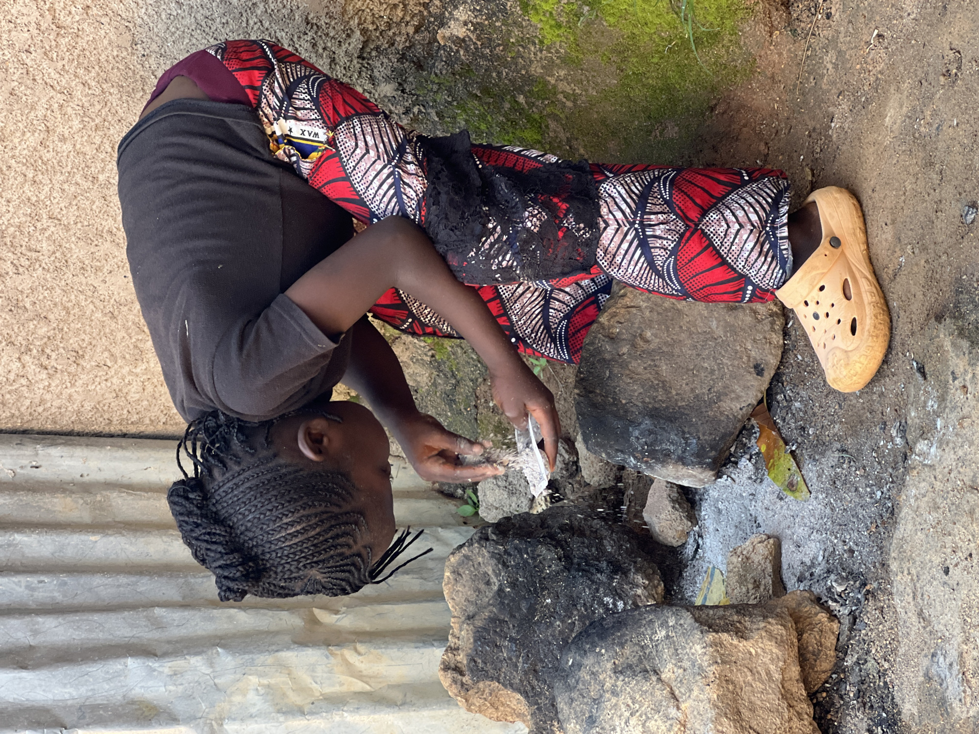 A young person with braided hair is bent over, holding a small white dish in their hands. They appear to be sprinkling something from the dish onto the ground near a fire pit constructed from large rocks. The person is wearing a dark t-shirt and brightly patterned trousers with red, white, and black designs, and an orange clog on their right foot. The background features a corrugated metal wall on the left and a textured, beige wall on the right, with moss growing on parts of it. The ground is dirt and gravel, with scattered ashes and stones, suggesting an outdoor cooking or fire-making area. The scene is set in a humble, likely domestic outdoor space, characteristic of a location in Jos, Nigeria.
